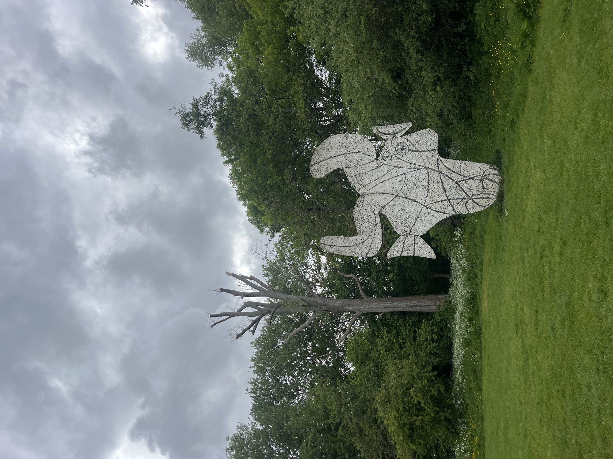 The image depicts a mosaic sculpture of a fantastical creature, possibly a bird, standing on a grassy lawn surrounded by trees. The sculpture is white with black lines and details. Behind the sculpture is a tall, leafless tree and a backdrop of green trees under a cloudy sky. The photograph appears to be taken in a park or garden setting. It is likely located in Amsterdam, Netherlands, given the context. There are no people visible in the image.
