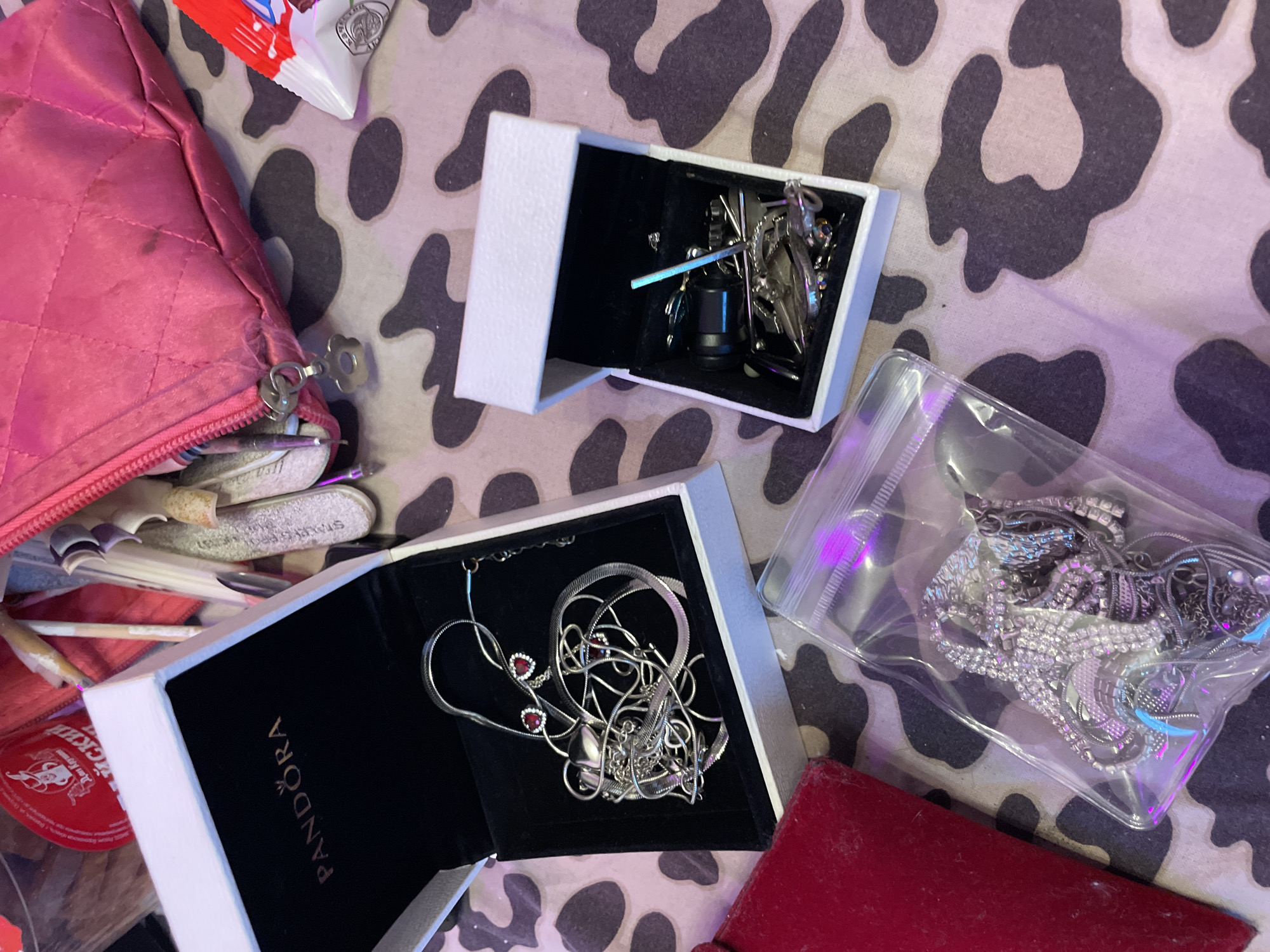 The image displays a collection of jewelry and cosmetic items laid out on a patterned surface, likely a table or bedspread, with a leopard-print design in shades of pink and gray. In the upper left quadrant, a red circular object with text in Cyrillic script is visible. To its right, a pink quilted cosmetic bag is open, revealing various items, possibly makeup brushes and nail files, protruding from it.
Central to the image is an open white Pandora jewelry box, containing a tangled silver necklace with two heart-shaped red stones and a delicate chain. To the right of the Pandora box is another smaller white box, also open, holding a variety of metallic accessories, including what appear to be earrings or charms.
In the lower portion of the image, a clear plastic bag is filled with an assortment of silver-toned jewelry, including necklaces and possibly bracelets. A red rectangular object, possibly a book or case, is positioned to the left of the plastic bag. The lighting in the scene appears to be artificial, with a purplish hue cast on the plastic bag and some of the jewelry, suggesting the presence of colored lights or a specific photographic setup.
No specific details are discernible that would place this scene in the city of Ladozhskoe Ozero, Russia. The image captures a static arrangement of personal belongings, with no identifiable individuals present or actions taking place.