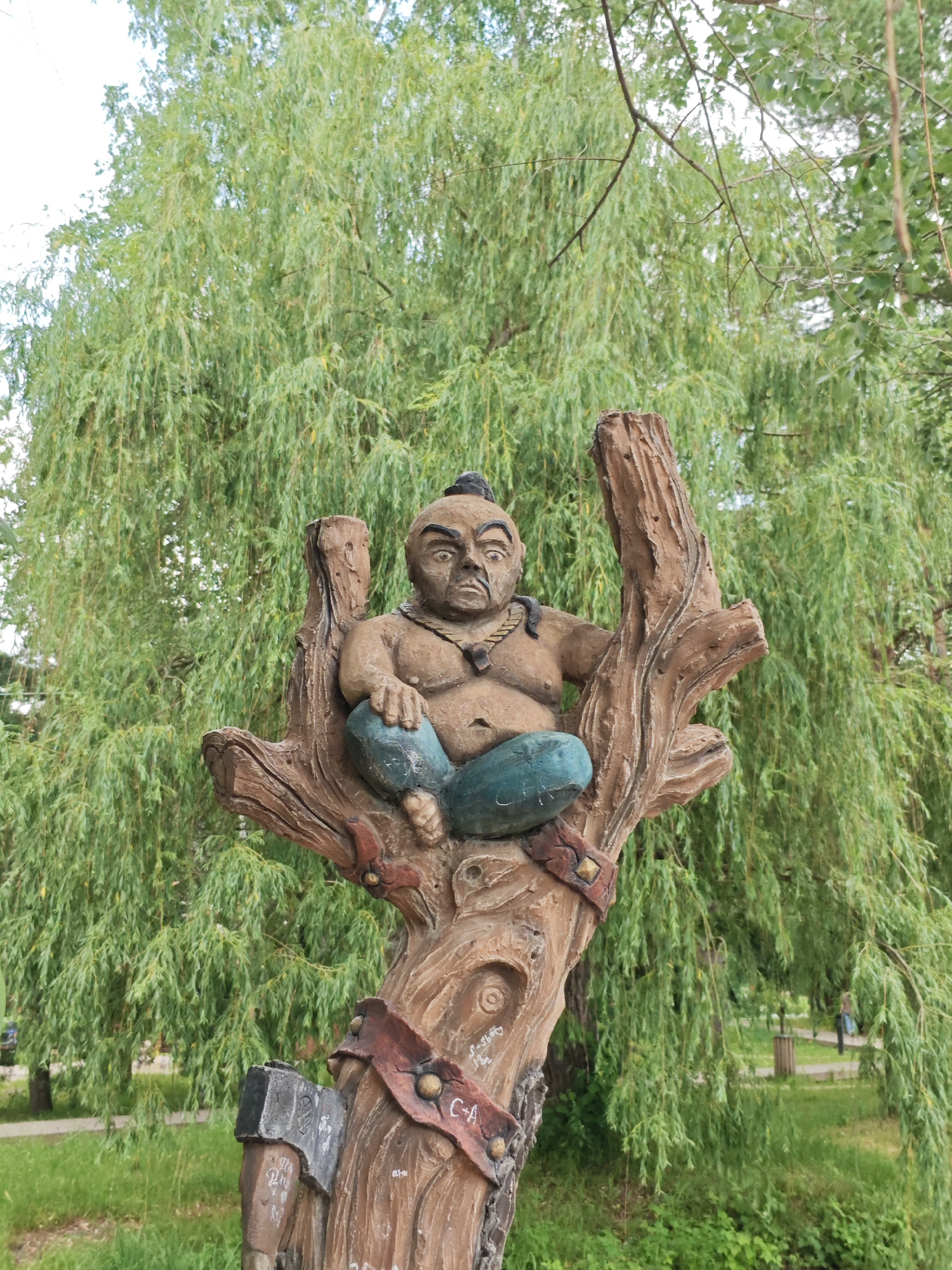 A carved wooden sculpture of a stout, seated figure with dark hair and a necklace is positioned in the fork of a tree-like wooden structure. The figure wears teal-colored pants and appears to be in a meditative pose. The sculpture is set against a backdrop of lush green weeping willow trees. The base of the wooden structure features an attached axe. The scene is outdoors, in what appears to be a park or green space. The location is identified as the city of Cherkessk, Russia. No people are clearly interacting with the sculpture.