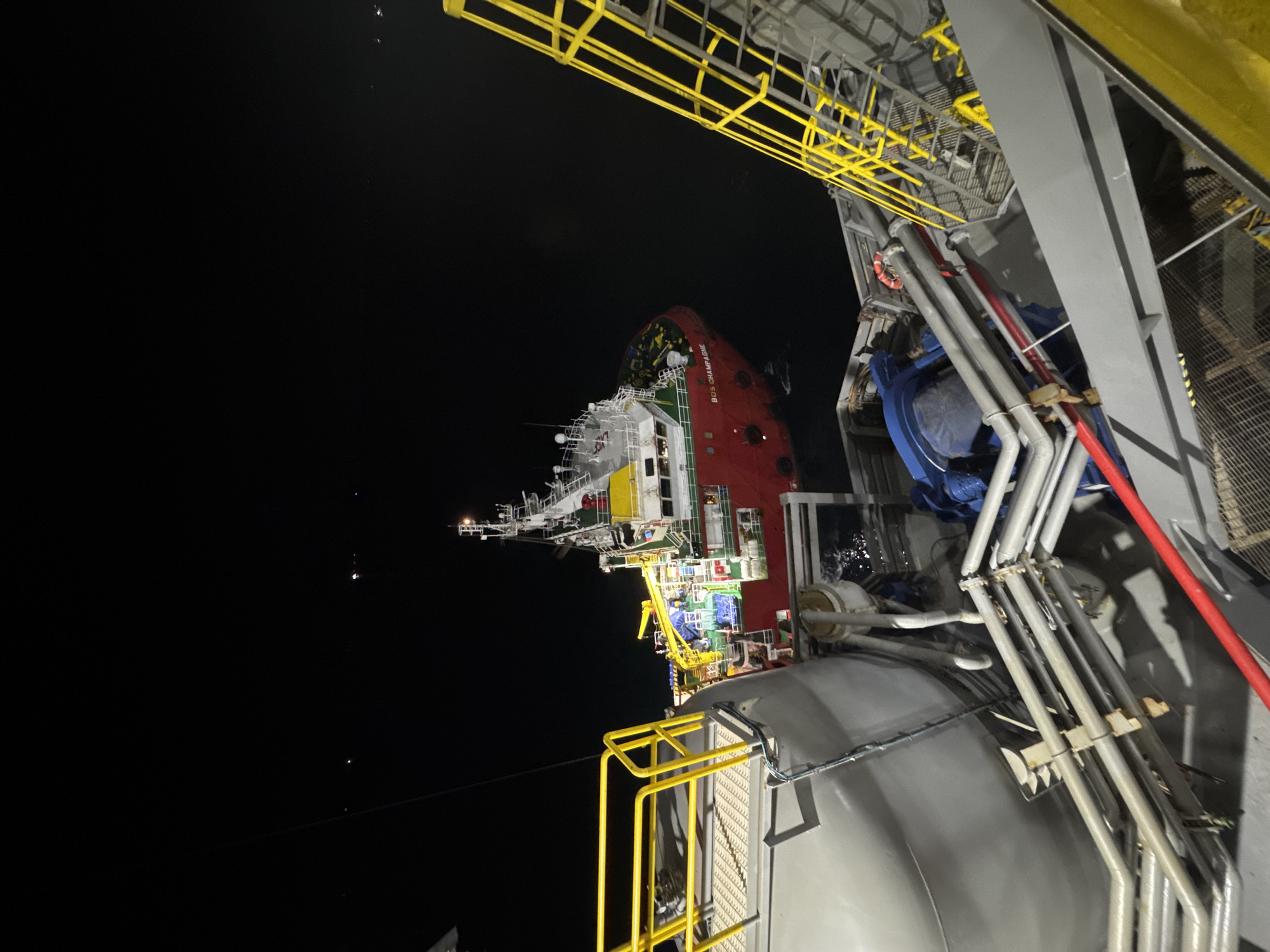 A nighttime, aerial view captures a large, red vessel with white superstructure, identified by the visible text "BOS CHAMPAGNE" on its side, docked or moored near a grey industrial structure. The vessel appears to be a ship, likely a supply or support vessel, given its prominent deck equipment and the cranes visible. The industrial structure features extensive yellow metal grating and railings, suggesting a platform, dock, or another vessel. Several pipes, some grey and some red, are visible along the side of the industrial structure, suggesting an operational or active environment. The scene is illuminated by artificial lights, creating stark contrasts between the illuminated areas and the deep black night sky. The water is not clearly visible, but the reflections on the side of the industrial structure hint at the presence of liquid, possibly water or a spill. The overall impression is one of industrial activity at night, likely in a port or offshore facility. The sky is dark, indicating nighttime, and no specific weather conditions are apparent. The image is oriented such that the horizon is vertical.