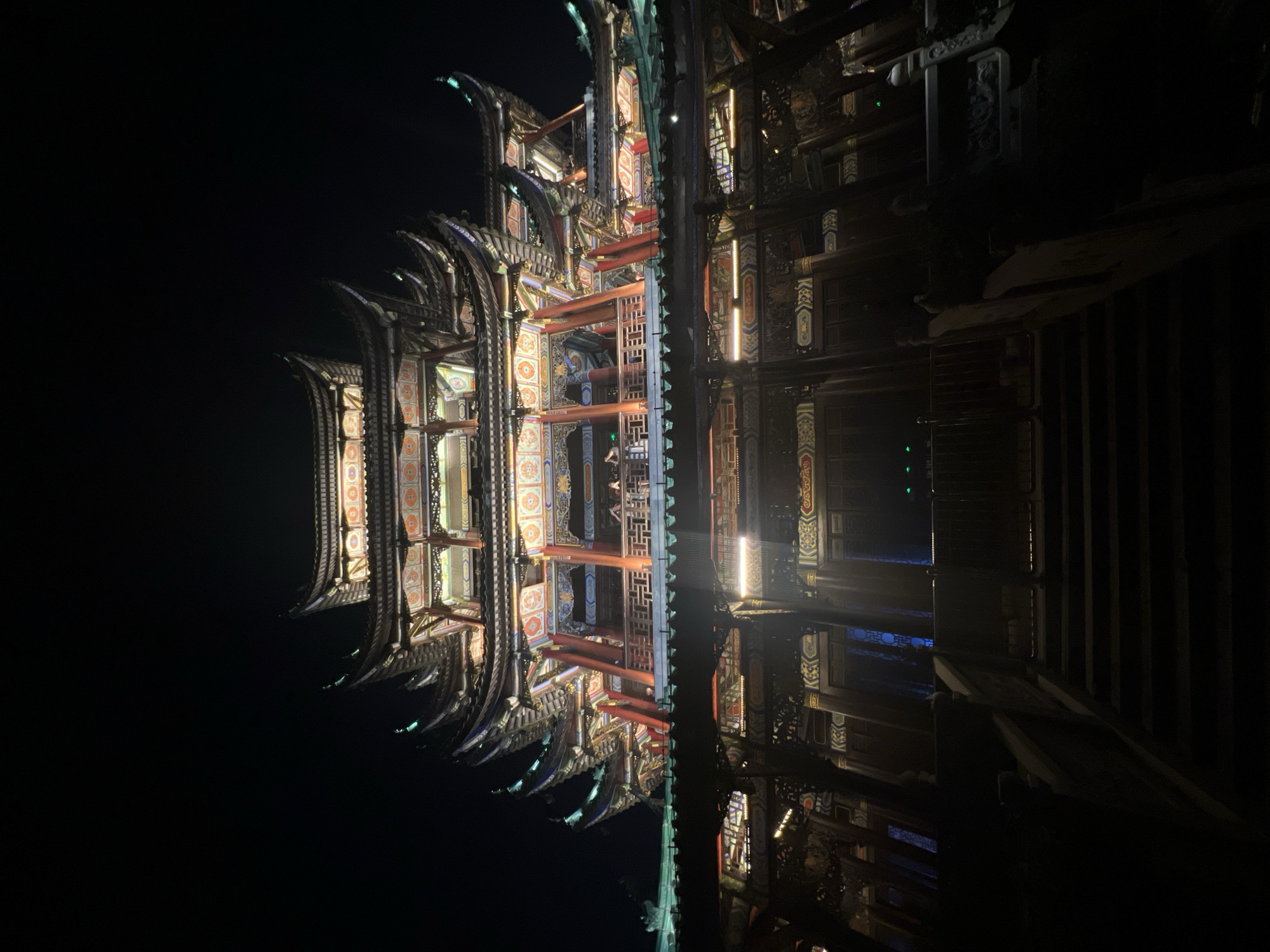The image displays a multi-tiered, elaborately decorated traditional Chinese building at night. The structure features multiple curved roofs, ornate carvings, and vibrant colors, including red, gold, and blue, illuminated by artificial lighting. The building is viewed from a low angle, looking upwards from a dark, possibly stone staircase in the foreground. Several individuals can be seen on a balcony or walkway on one of the upper levels, silhouetted against the lights. The overall scene suggests a cultural or historical site, likely a temple or a significant architectural landmark, in the city of Liangjiang, China. The sky is dark, indicating it is nighttime.