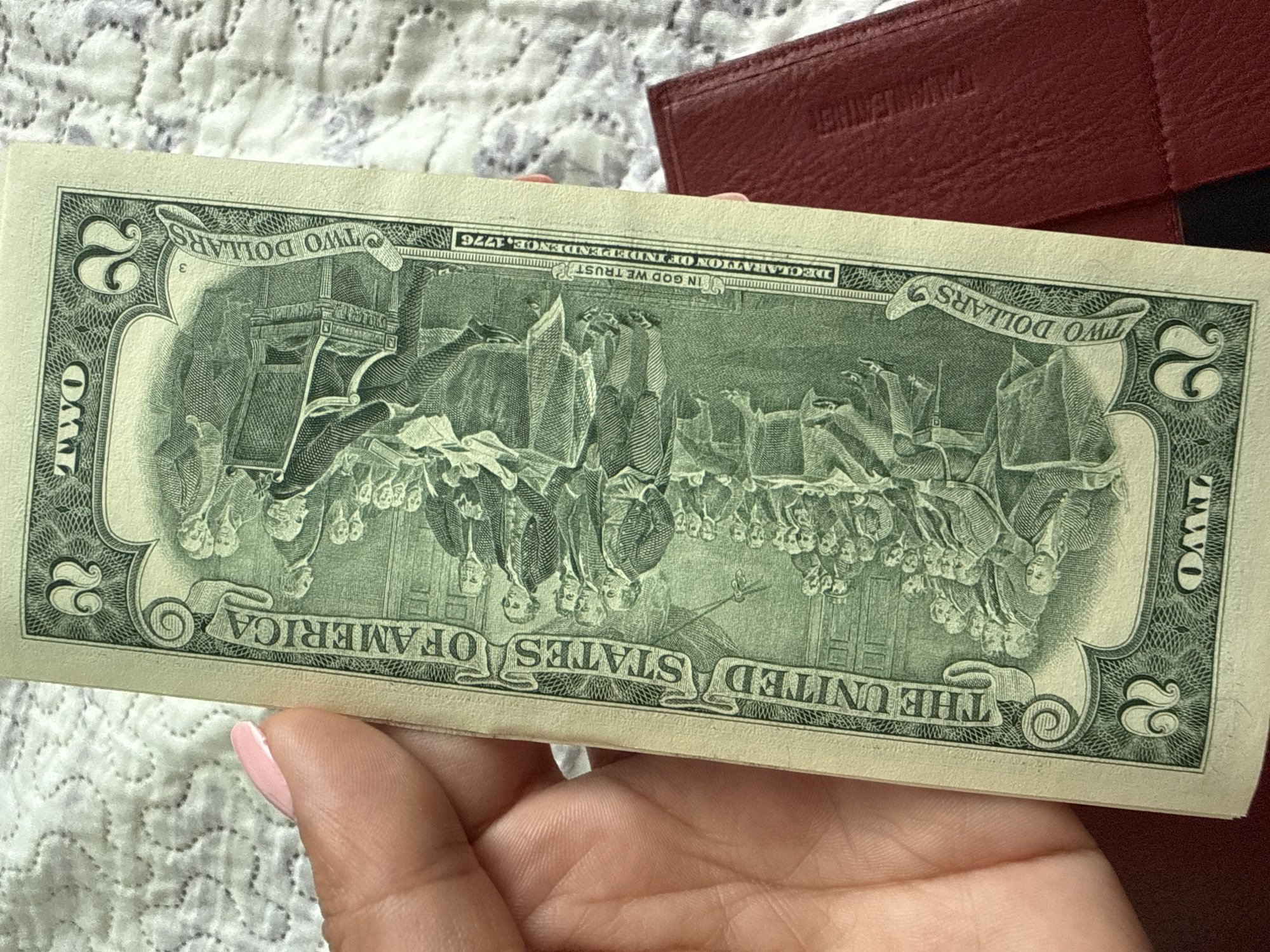 A close-up, vertical, eye-level shot shows the back of a United States two-dollar bill being held by a person's left hand. The bill features a depiction of the "Declaration of Independence" with many figures in historical attire. Text on the bill reads "THE UNITED STATES OF AMERICA," "TWO DOLLARS," "IN GOD WE TRUST," and "DECLARATION OF INDEPENDENCE, 1776." The bill is positioned over a textured surface, possibly a blanket, with a red leather object partially visible in the lower right quadrant. The hand holding the bill has light-colored skin and a light pink fingernail polish. The image does not contain any elements that can be used to determine the location in the city of Ekzarh Antimovo, Bulgaria.
