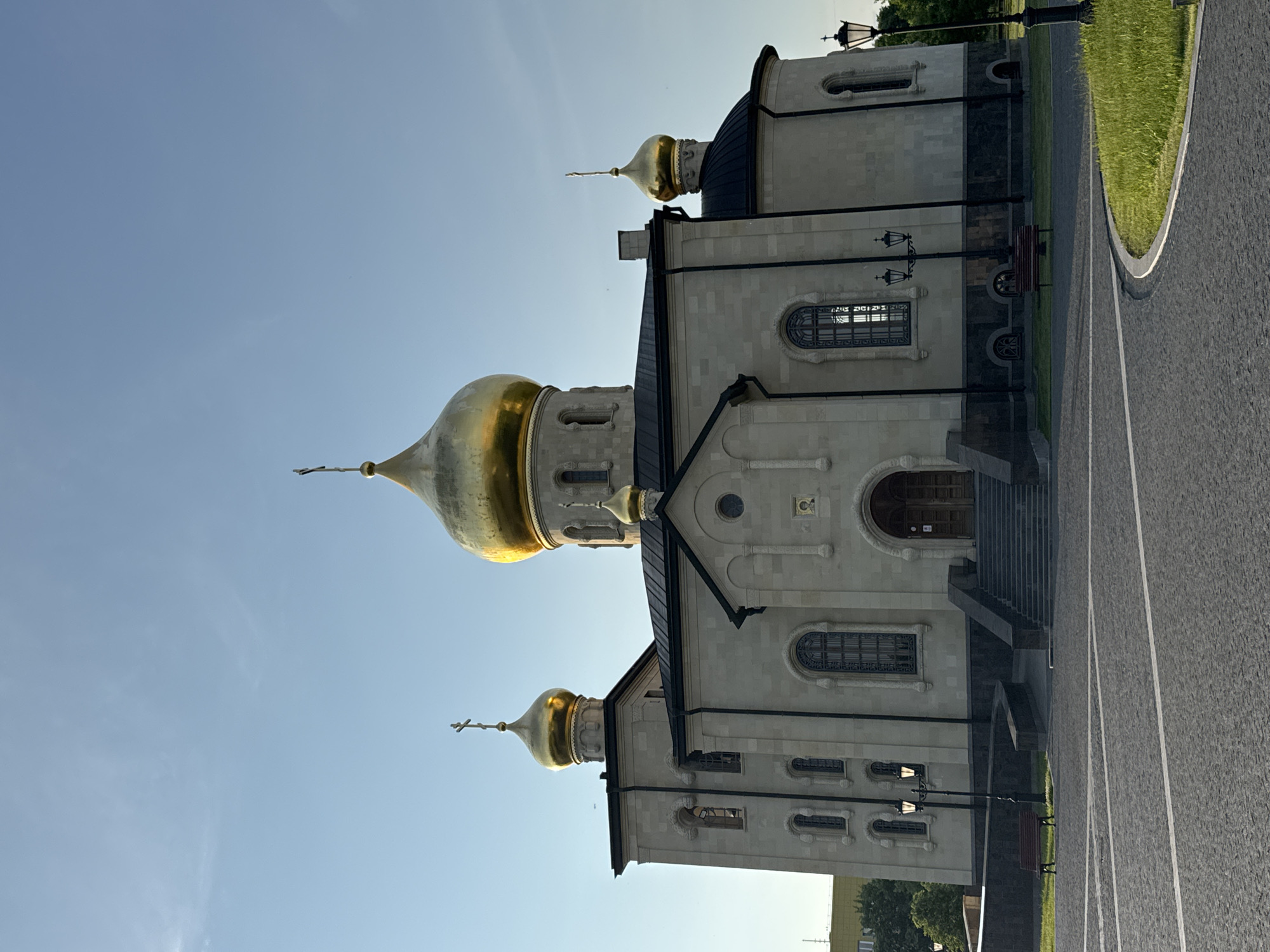 The image displays an Orthodox church with golden domes against a clear blue sky. The church is constructed of light-colored stone and features a grand entrance with stairs leading up to wooden doors. Several windows, some with intricate designs, are visible on the facade. A prominent golden dome sits at the center of the structure, flanked by smaller golden domes on towers and on the sides of the building. The church is situated in an open, paved area that appears to be a public square, with some greenery and benches visible. Streetlights are also present around the church. The overall scene suggests a serene and well-maintained religious site. This is the Church of the Resurrection of Christ, located in the city of Kirishi, Russia.