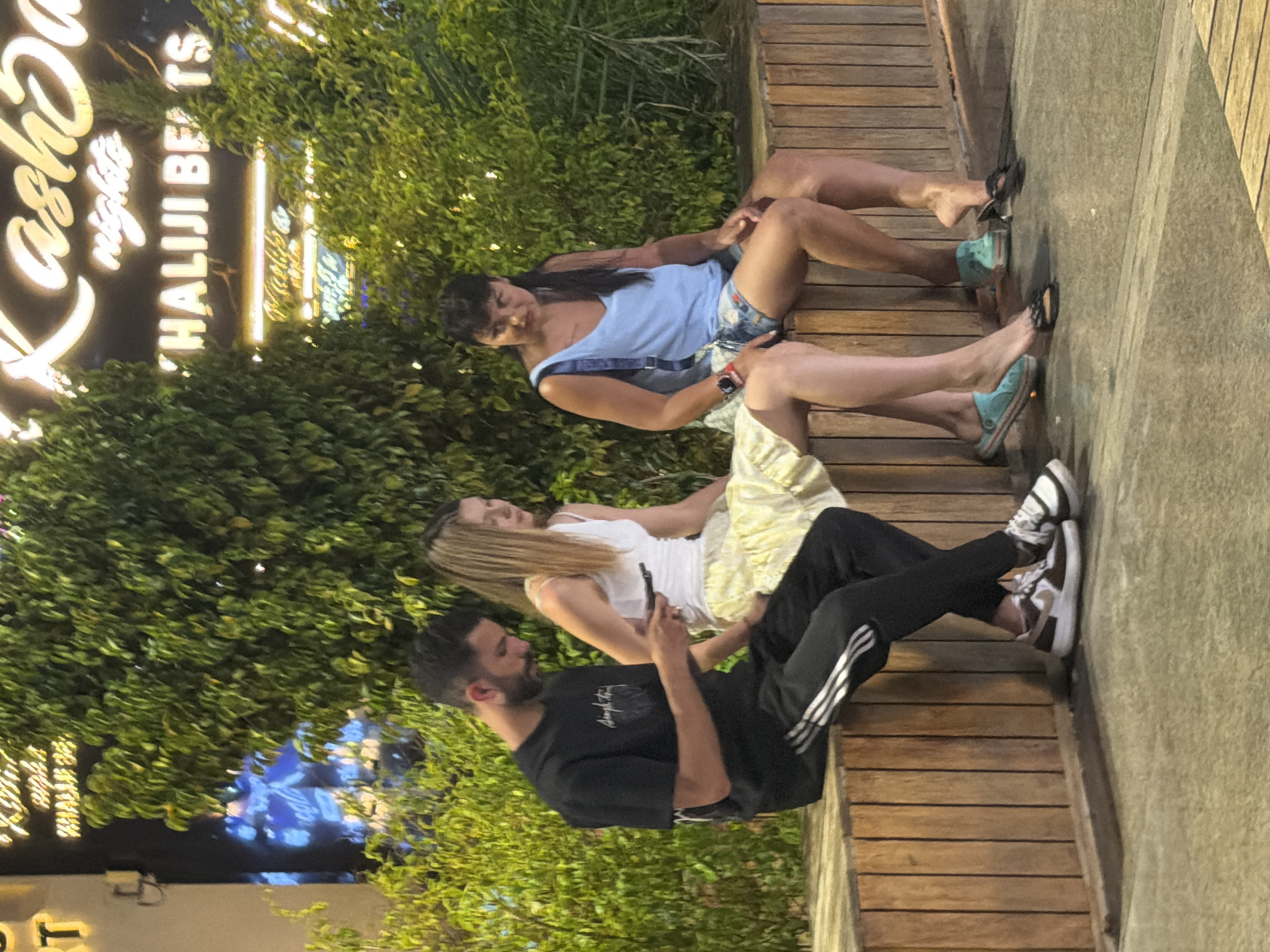 Three individuals, two women and one man, are seated on a wooden bench in front of dense green foliage. The man on the left is wearing a black t-shirt and black track pants with white stripes, and is looking down at a mobile phone in his hand. The woman in the middle is wearing a white tank top and a yellow patterned skirt, and is looking forward. The woman on the right is wearing a light blue sleeveless top and denim shorts, and is also looking forward. Behind them, a neon sign displays "Kashsa" and "HALIJI BEATS," indicating a night venue, possibly in Sharm El Sheikh, Egypt, known for its entertainment districts. The ground in front of the bench appears to be paved.