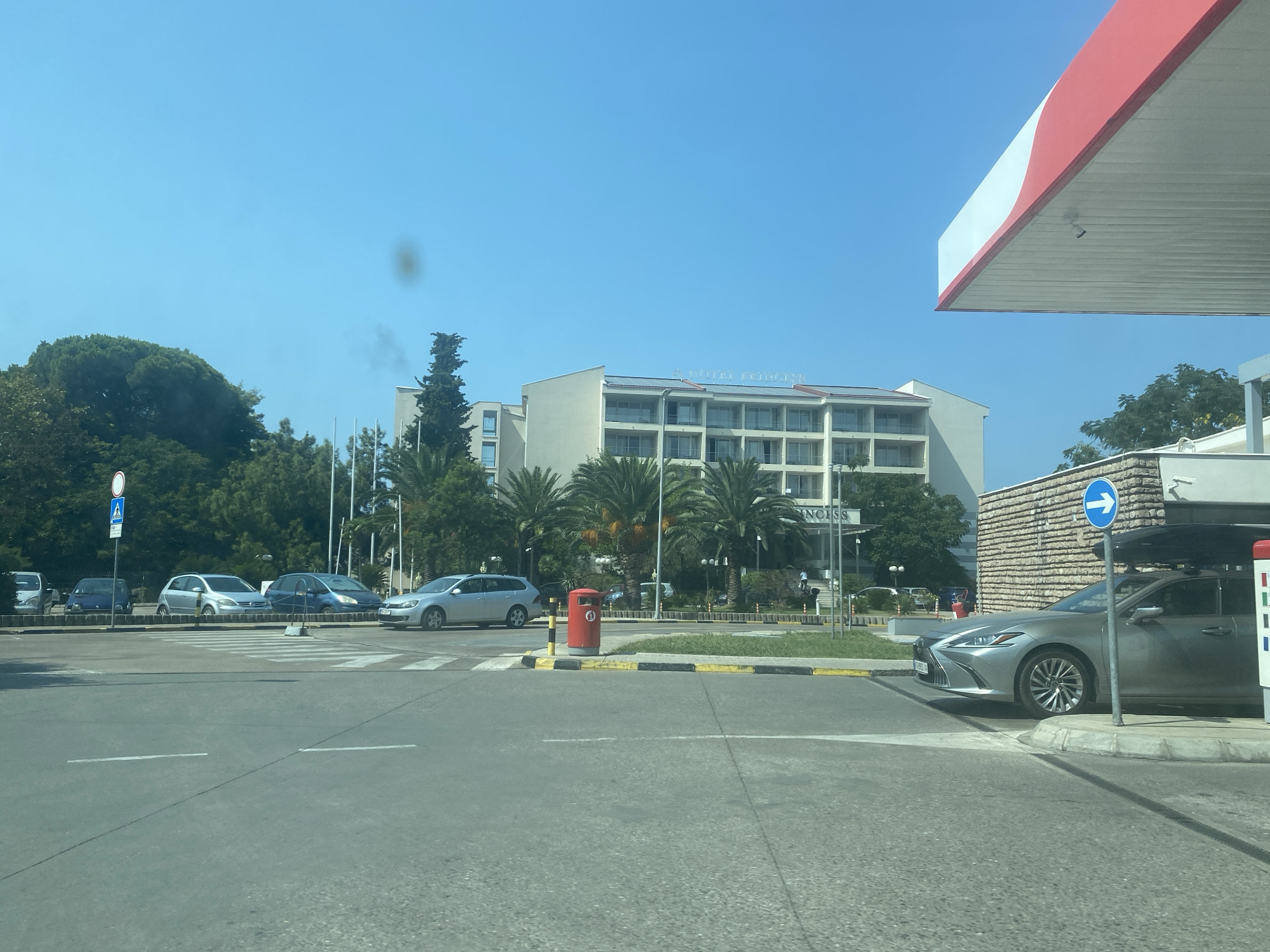 A bright, sunny day is depicted in this scene, likely in the city of Bar, Montenegro, near a gas station. The foreground shows a paved road with white lane markings. To the right, the red and white canopy of a gas station overhangs the scene, with a surveillance camera visible underneath. A silver sedan is parked to the right, near a stone-faced building. In the mid-ground, a crosswalk is visible, with several cars parked behind it. Among the parked cars are a silver hatchback and a blue sedan. A red trash bin stands next to the crosswalk. In the background, a large, multi-story hotel with balconies and palm trees in front of it is prominent. The hotel has a sign that reads "PRINCESS". Trees and other buildings are also visible in the background. The sky is clear and blue.