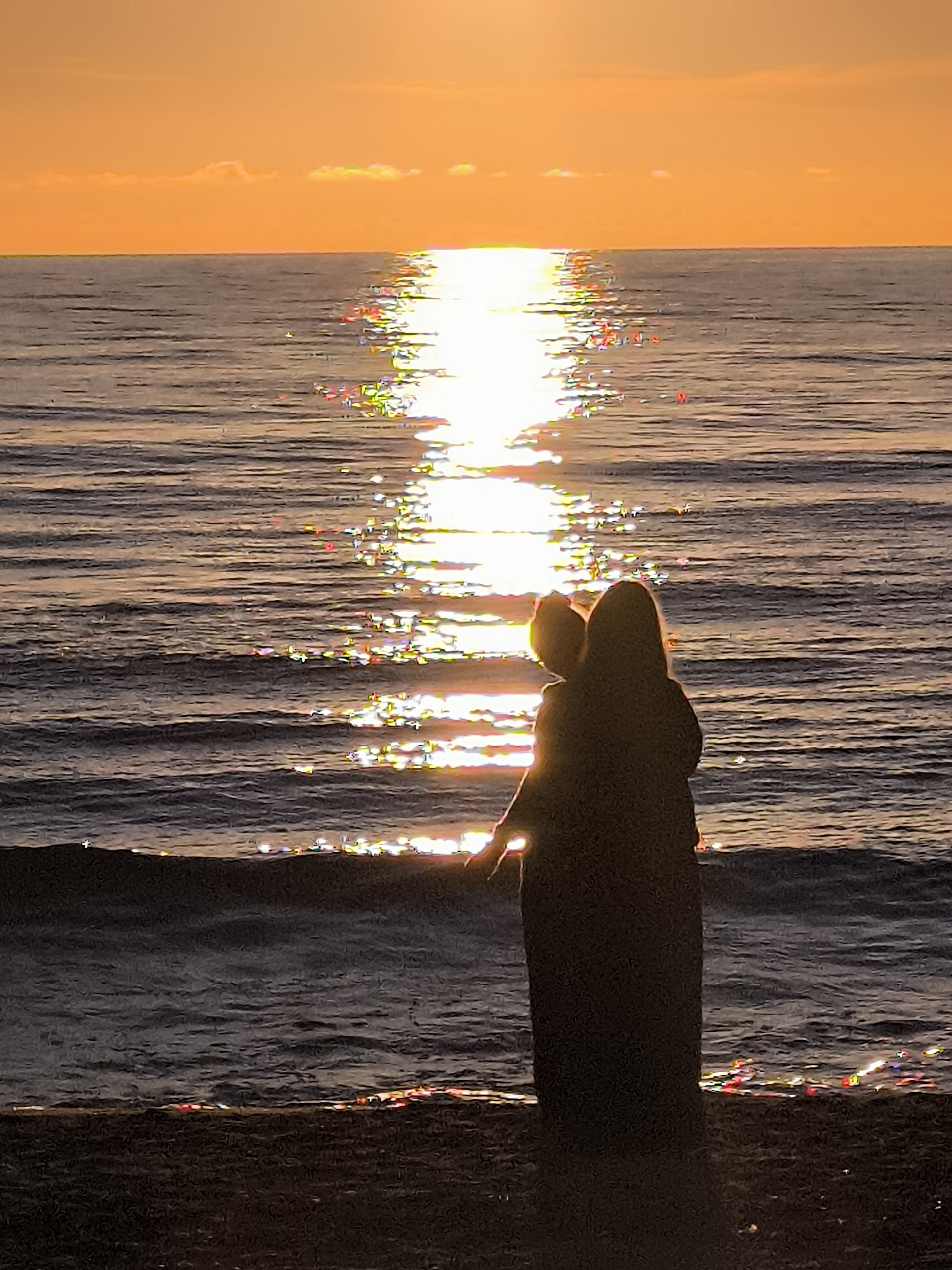 The image depicts a serene sunset over the Baltic Sea in Klaipėda, Lithuania. In the foreground, silhouetted against the vibrant orange and yellow hues of the sky and the shimmering reflection of the sun on the water, is a person holding a child. They stand on a dark, sandy beach, facing the expansive ocean. The water is calm with gentle ripples, and the sun's reflection creates a bright, broken path across its surface, interspersed with colorful glints. The horizon is clearly defined, separating the sea from the sky, where a few wispy clouds are visible. The overall mood is peaceful and contemplative, capturing a moment of shared experience between a parent and child as they witness the natural spectacle.
