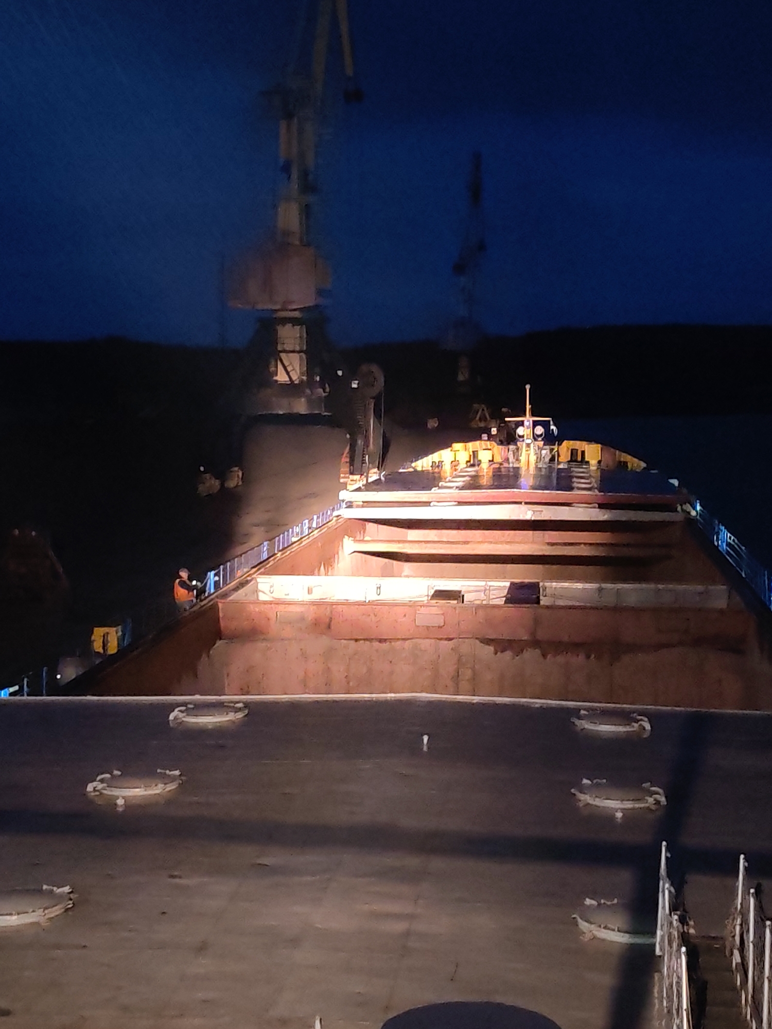 The image depicts a large cargo ship being loaded at night. A crane is positioned over the ship's hold, presumably loading or unloading cargo. A person wearing a high-visibility vest stands near the ship's railing. The scene appears to be located in a port or harbor, likely in the vicinity of Shalsky, Russia, given the prompt's specifications. The environment is dark, illuminated by artificial lights.
