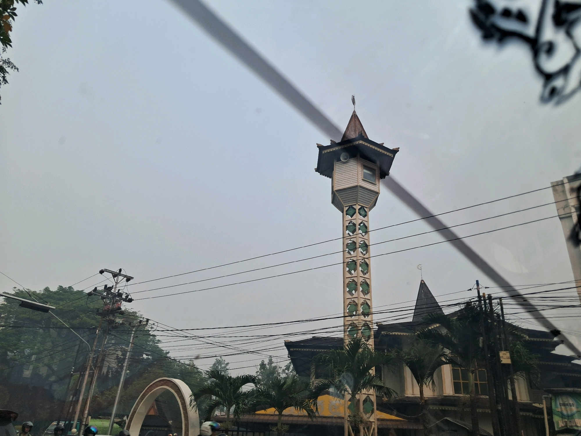 This image captures a street view in Pontianak, Indonesia, under an overcast sky. The focal point is a distinctive minaret with a white octagonal structure adorned with green diamond-shaped windows. The minaret stands tall next to a traditional building with a dark roof and arched windows. Several power lines crisscross the sky, casting shadows on the scene. In the foreground, palm trees and lush green foliage are visible, with some vehicles and people on motorcycles suggesting street activity. A semicircular archway is also present, hinting at an entrance or decorative structure. The overall impression is a snapshot of daily life in a tropical urban environment.