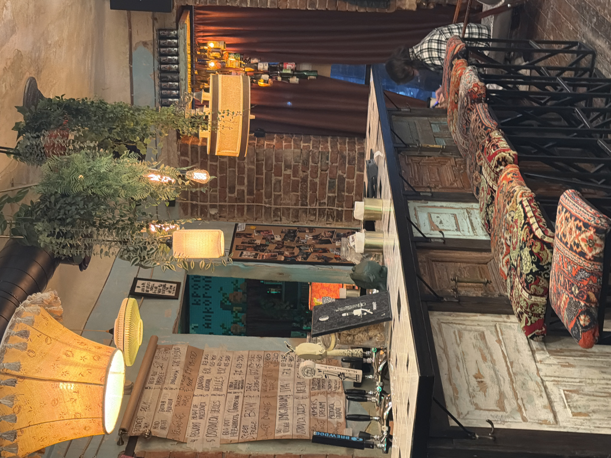 The image captures the interior of a bar or pub in Rostov-on-Don, Russia, displaying a rustic and eclectic aesthetic. A person in a plaid shirt is visible in the background, near a bar counter lined with stools adorned with patterned fabrics. The bar area features vintage or repurposed doors, arranged vertically, as part of the decor. Above, there are dark metal structures, possibly serving as decorative or structural elements.
A menu board listing various beers with corresponding descriptions and prices is positioned next to beer taps. A variety of light fixtures, including a lampshade with fringed edges and exposed light bulbs, illuminate the scene, adding to the ambiance.
The environment is cluttered yet designed, with hanging plants, decorative items, and a photograph collage adding visual interest.