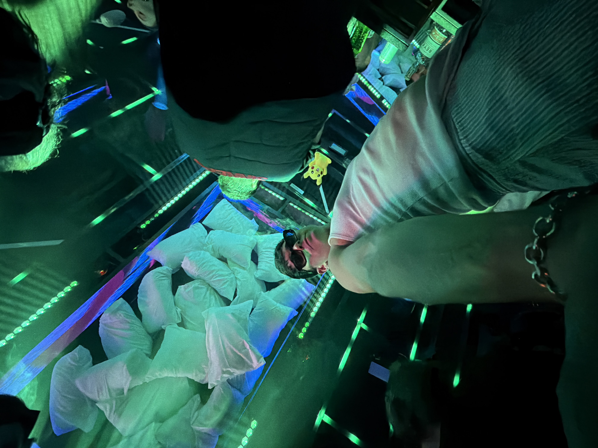 The image appears to be taken inside a "Pillow Room", an immersive art installation in the city of Sofia, Bulgaria. The Pillow Room consists of a mirrored enclosure with pillows scattered on the floor, illuminated with green and blue LED lights. The perspective is from above, looking down into the room. At least three people are visible: one wearing a grey shirt and chain, another with sunglasses and a dark-colored shirt, and another behind them. There is a small Pikachu toy in the middle. A bottle and glass can be seen at the upper part of the image.