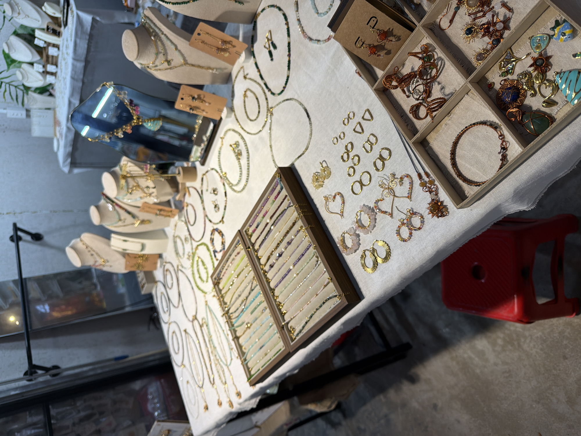 The image shows a table displaying jewelry, likely at a market or fair. The items include necklaces, bracelets, and earrings. The jewelry is made from various materials, including beads, metal, and gemstones. A wooden display case holds several bracelets. A red plastic stool is near the table. The environment suggests an indoor setting, possibly in Đà Nẵng, Vietnam, though without further context, precise location is impossible to ascertain.