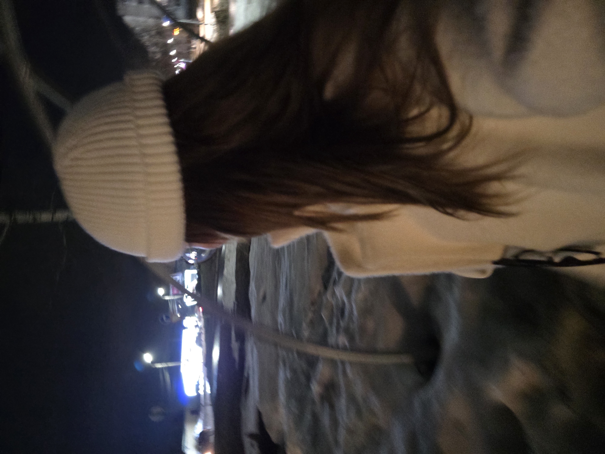 The image depicts a nighttime urban scene in Chaikovsky, Russia, under cold weather conditions, with patches of snow and ice visible on the ground. A person, likely female, is viewed from behind, occupying the foreground. This individual wears a white ribbed beanie hat and a light-colored coat, with long dark brown hair flowing beneath the hat. A slender metallic pole is present in the mid-ground, slightly to the left of the frame's center. The background is blurred, showing a street or intersection illuminated by multiple streetlights and vehicle headlights/taillights, indicating active traffic. Indistinct building structures are faintly visible in the far distance against a dark night sky.