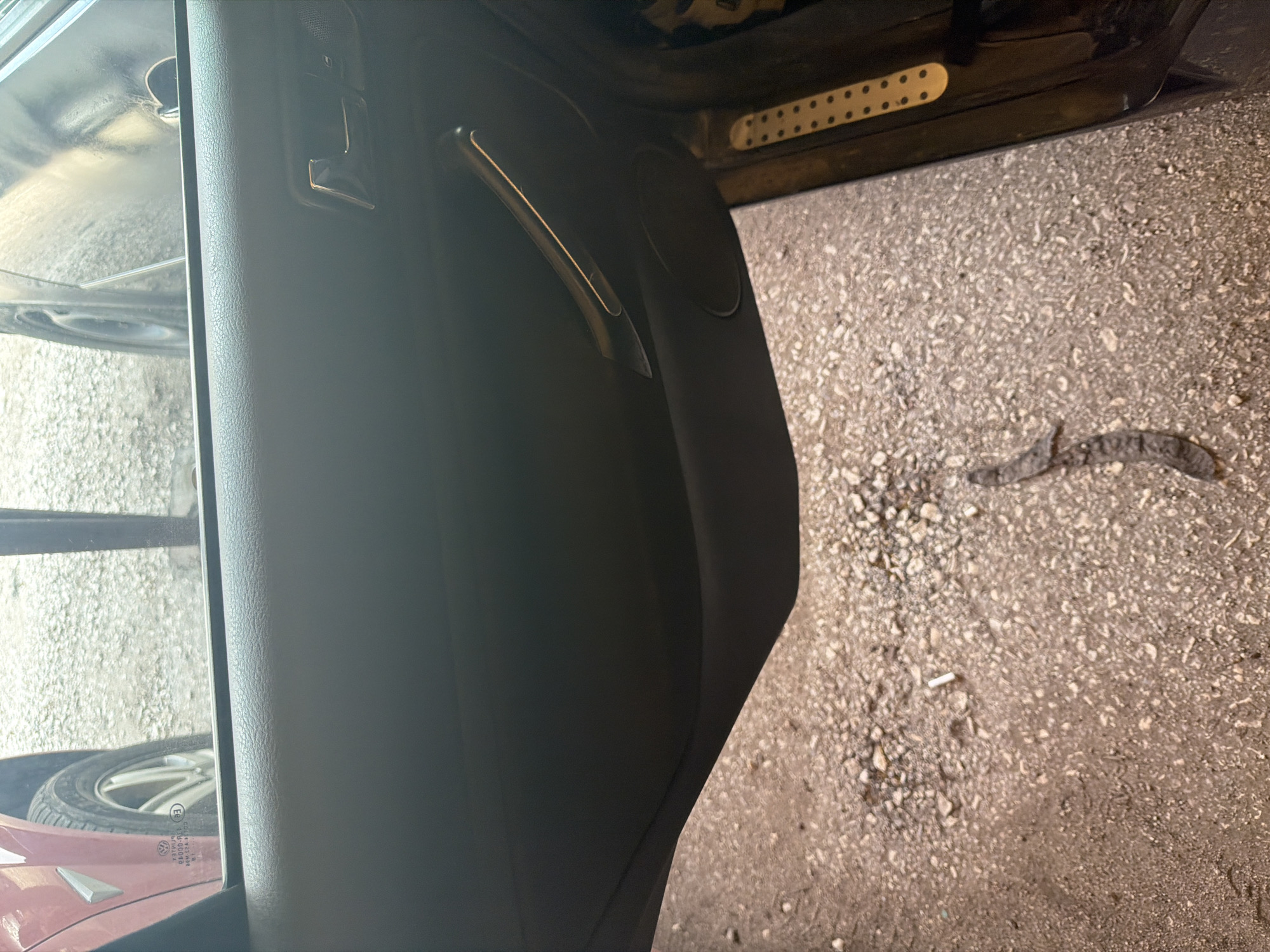 The image depicts a view from inside a vehicle, through an open car door, onto a concrete or asphalt ground surface. The vehicle door is black, with a silver handle. A cigarette butt lies on the ground. A snake, approximately 2-3 feet long, is also present on the ground surface. Through the car window, the tire and body of another vehicle (possibly a red Volkswagen) are visible. The location is undetermined. No people are visible.