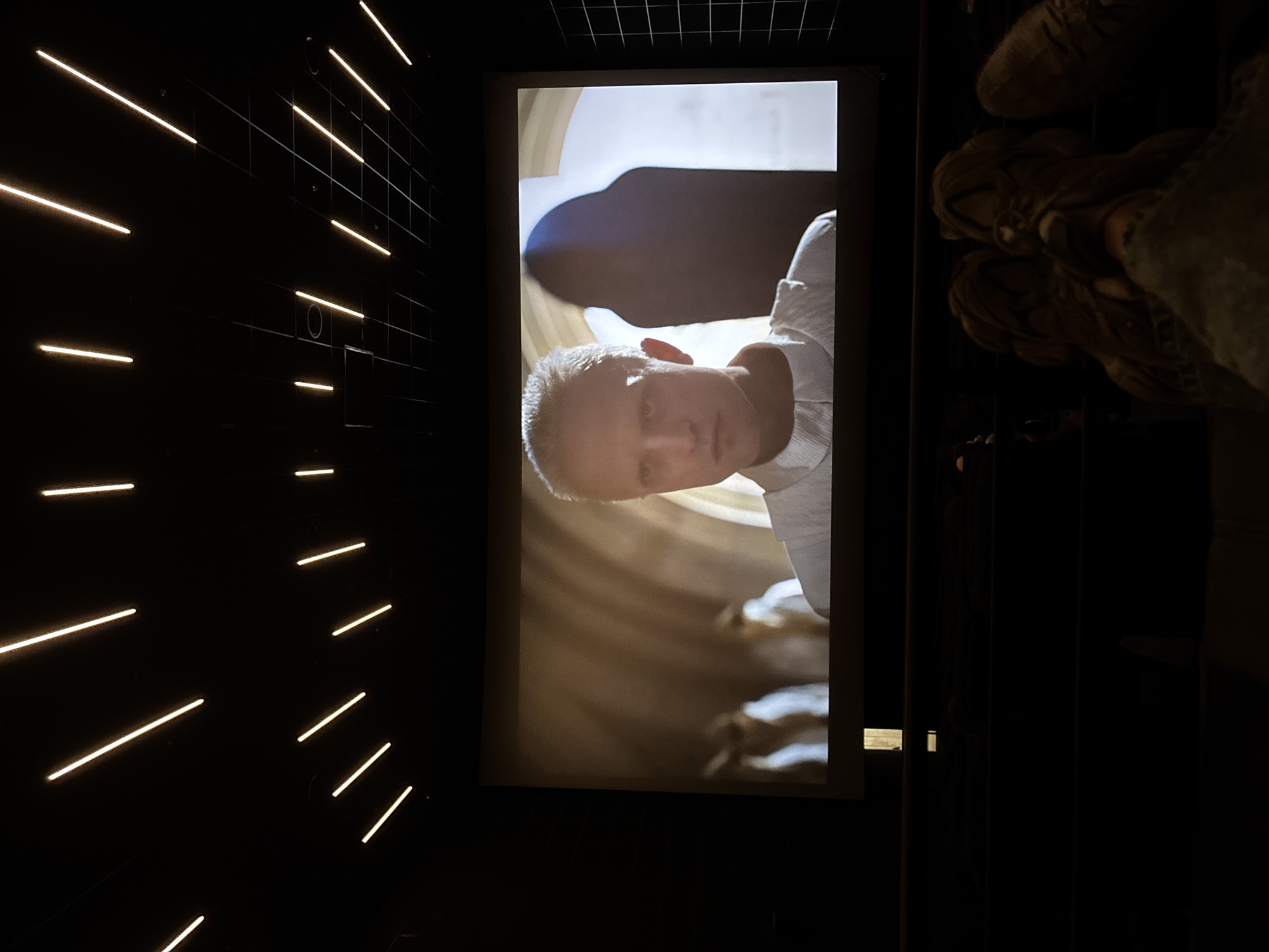 The image shows a person's face displayed on a screen within a dark room. The room appears to have a grid-like ceiling and a wall with horizontal light strips. The person on the screen has short, light-colored hair and is wearing a white shirt. The background on the screen suggests an interior space, possibly with architectural details. The overall environment is dim and modern, with the screen serving as the primary light source in that section of the room. I cannot verify the location of this image being in Kharkiv, Ukraine.