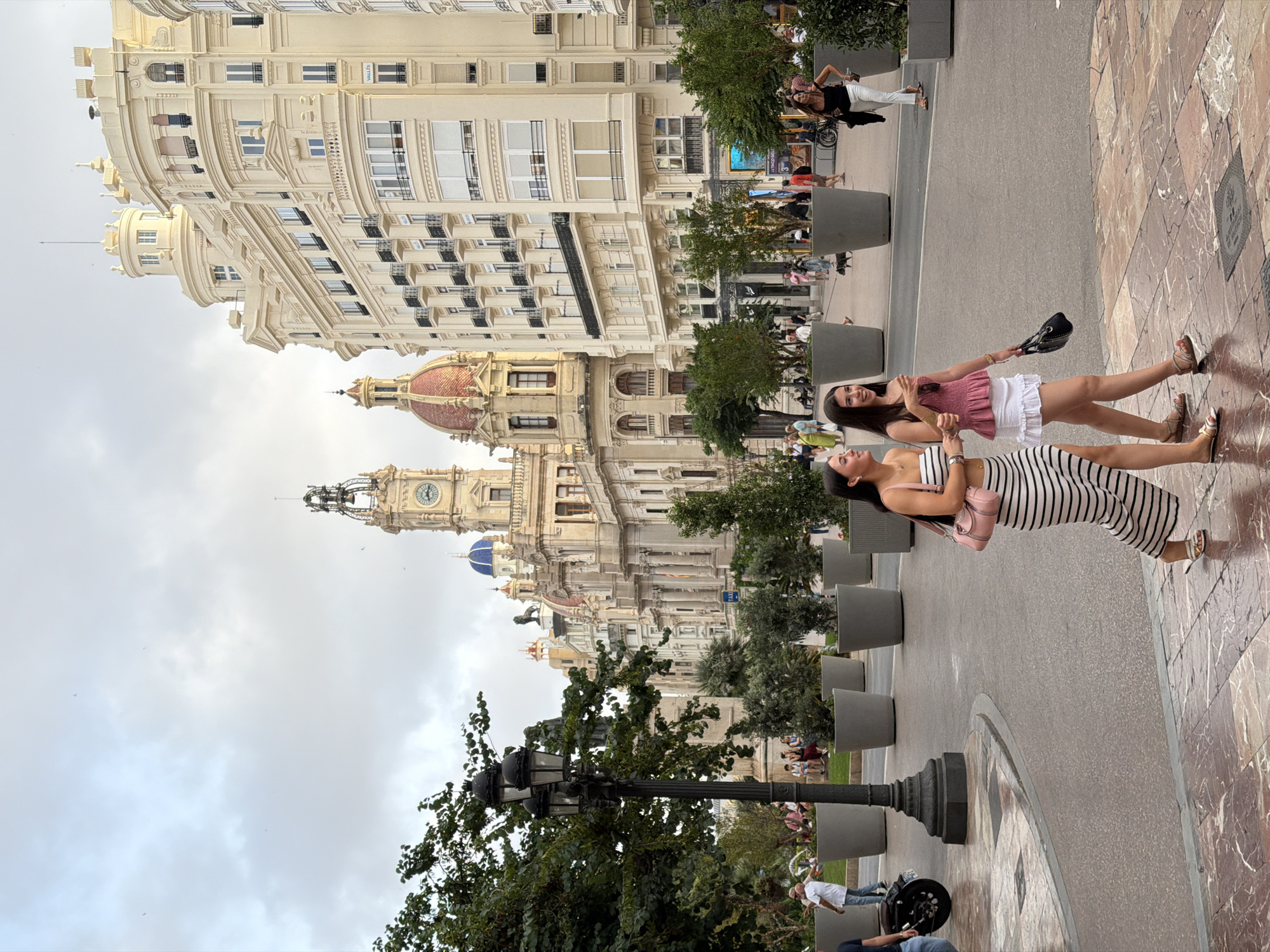 In a city square, two young women walk side-by-side across a cobblestone and marble patterned pavement. The woman on the left wears a black and white striped maxi dress and carries a pink handbag. The woman on the right wears a pink ruffled top and a white skirt, and carries a black handbag. In the background, a street lamp stands to the left, and large potted plants are arranged in rows. Several people can be seen in the distance, as well as ornate buildings with balconies, domes, and a clock tower. The sky is overcast.
