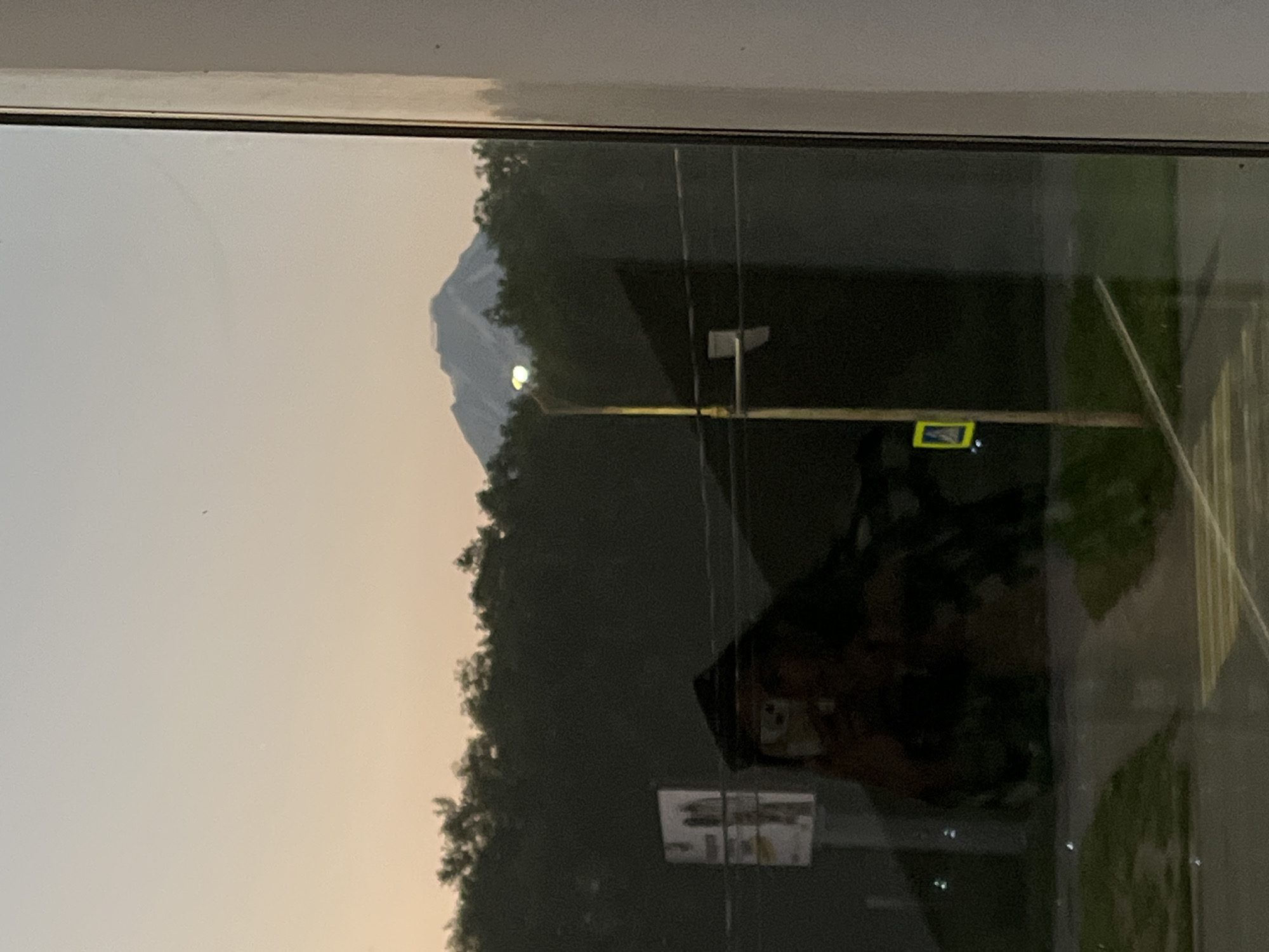A reflection in a window shows a distant mountain, possibly Mount Avachinsky or Mount Koryaksky, in Petropavlovsk-Kamchatsky, Russia. The sky is a gradient of pale orange to light blue, indicating either dawn or dusk. A dark line of trees forms a dense horizon below the sky. In the foreground, reflected in the glass, is a dark, indistinct shape that appears to be a person or a large object, possibly a coat or a silhouette. A street light with a bright yellow glow is visible, along with a yellow pedestrian crossing sign. The reflection also shows a portion of a road with white crossing lines and green grass. The overall scene is captured from an indoor perspective, looking outwards through a window frame.