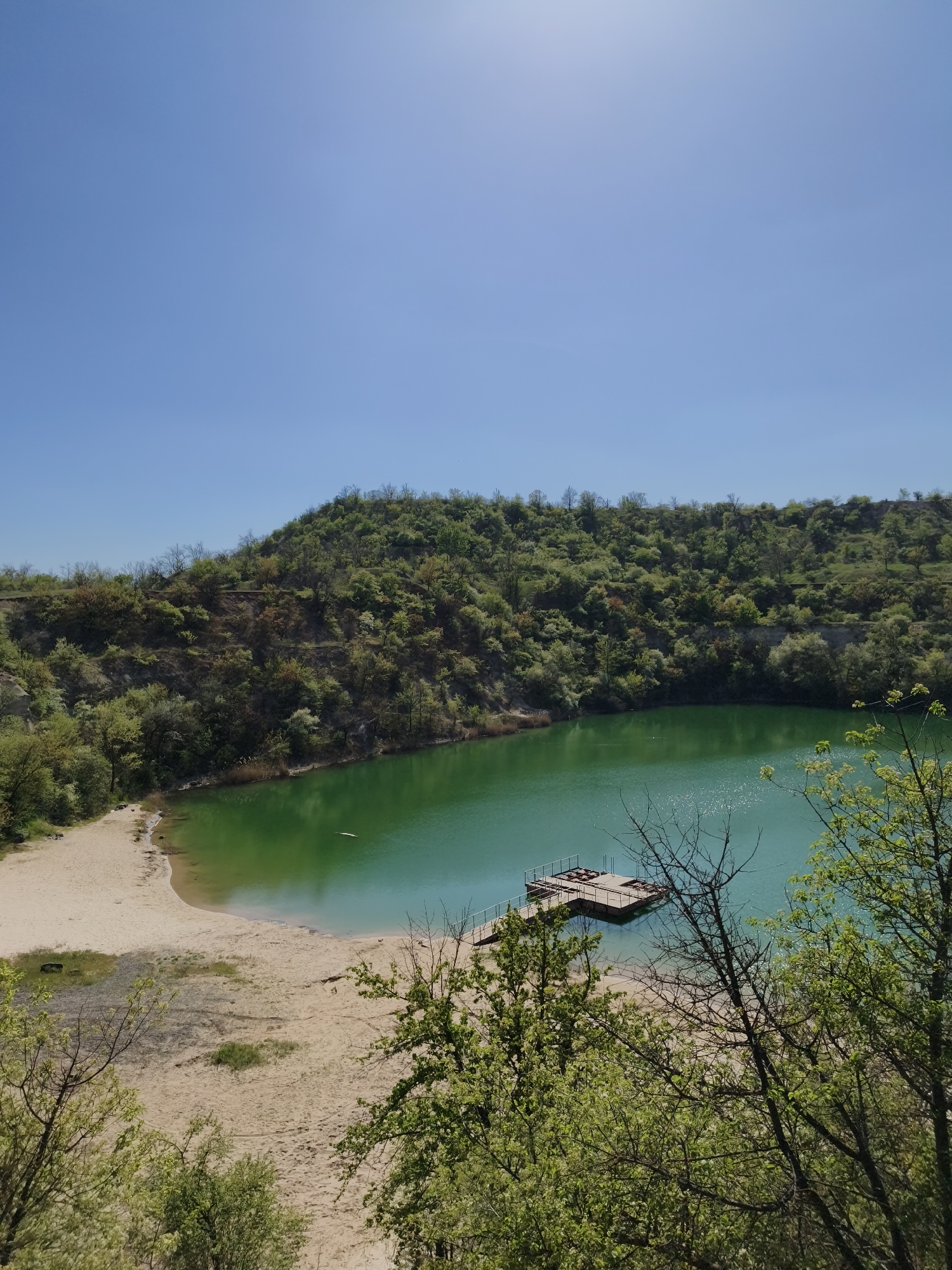 The image depicts a quarry lake located in the city of Kryvyi Rih, Ukraine. The lake has a bright green color and is surrounded by a sandy beach on one side and a hill covered with trees and vegetation on the other. In the water, there is a wooden platform or dock structure. The sky is clear and blue with the sun visible in the upper right corner of the frame. The foreground features trees and shrubs.