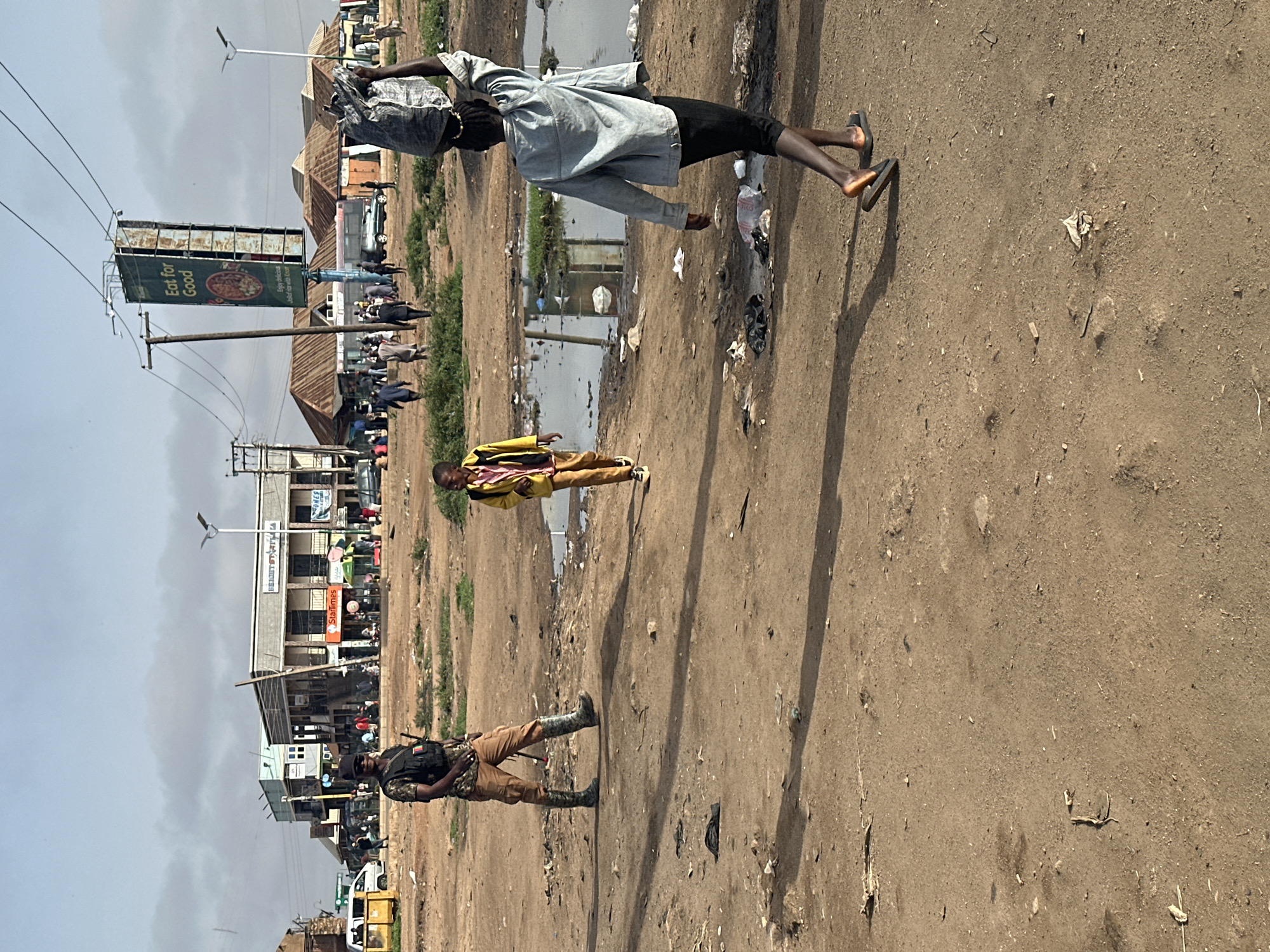 The image depicts an unpaved road in what appears to be a market area in Bukuru, Nigeria. The ground is dusty and contains debris.
Several individuals are visible:
* One person is carrying a heavy load on their head.
* Another person is wearing a yellow jacket.
* A third person is wearing a military-style uniform and boots.
In the background, there are several buildings and a billboard that says "Eat for Good." Other people are gathered in the area, suggesting an active marketplace. Puddles of water can be seen on the ground.