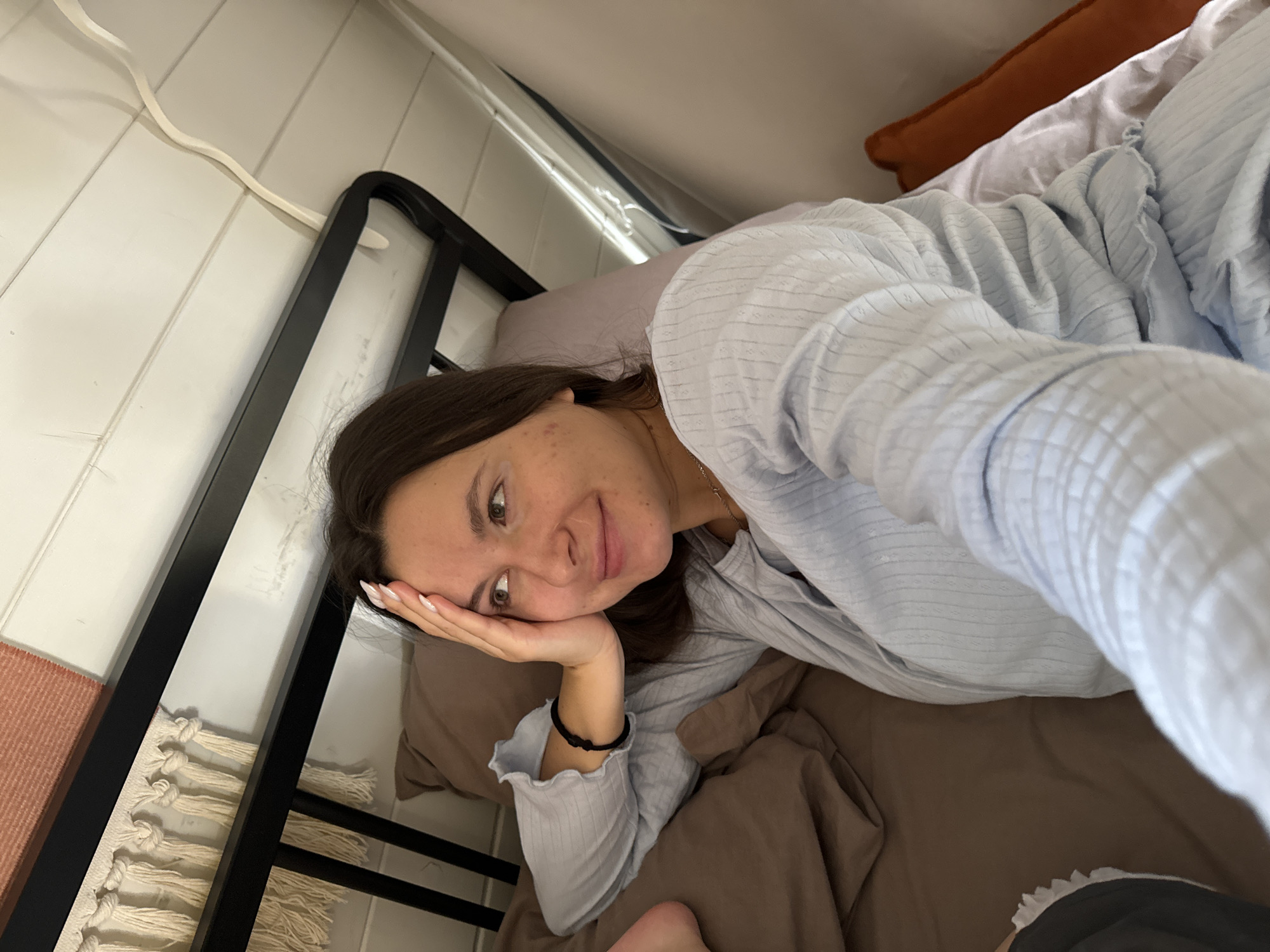 The image shows a woman lying in bed, appearing to take a selfie. She is resting her head on her hand, with a relaxed expression. The bed has brown sheets and pillows. The bed frame is black metal. The background includes a white, textured wall and a glimpse of an orange pillow. The location is Литвиново, Russia.