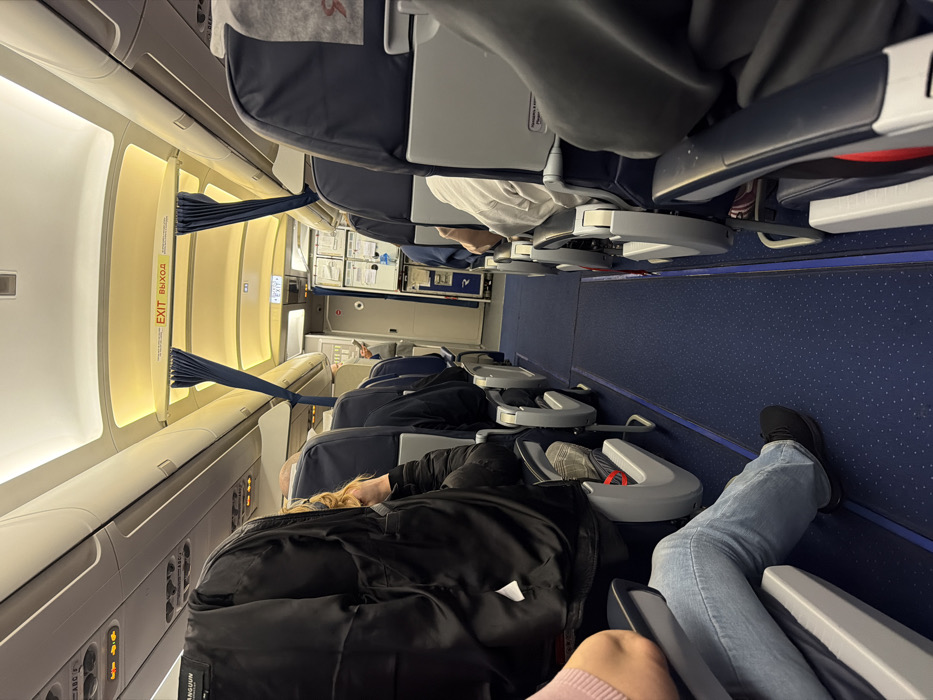 This image depicts the interior cabin of a commercial aircraft, presumably at Емельяново (аэропорт), Russia. The perspective is from a passenger seat, looking forward down the main aisle towards the front of the aircraft.
**Environment:**
The aircraft cabin features rows of blue and gray passenger seats arranged in a typical 3-3 or 2-2 configuration (specific number per row not fully visible). A narrow blue carpeted aisle runs down the center. Overhead bins are closed and illuminated by warm white LED lighting, which also illuminates the ceiling. An "EXIT ВЫХОД" sign, displayed in both English and Russian, is centrally positioned above the aisle in the forward section of the cabin. Dark blue curtains are pulled back on either side of this exit sign. Further forward, a galley or cabin crew station is visible, containing various panels and equipment.
**People:**
Multiple passengers are seated throughout the cabin. In the immediate foreground on the left, a person is seated, wearing light blue jeans and black athletic shoes. Their right arm, covered by a light pink long-sleeved garment, rests on the armrest. The head and torso of this individual are out of frame. The seat directly in front of this person is occupied by an individual in a black jacket, their head partially visible with blonde hair. Further down the left aisle, several other passengers are visible, primarily from the back of their heads and shoulders, occupying seats. On the right side of the aisle, a passenger is seated in the foreground, primarily visible from their right arm and side, wearing a light gray long-sleeved shirt and dark trousers. Other passengers are visible further down the right aisle, similarly seated. No clear active interactions between passengers are discernable.
**Objects:**
Identifiable objects include passenger seats with blue fabric and gray plastic frames, overhead bins, illuminated ceiling panels, the "EXIT ВЫХОД" sign, blue curtains, and the blue speckled carpet. The seatbacks have literature pockets and a small flip-down tray table (not in use). A logo, possibly "S7 Airlines," is faintly visible on the headrest cover of one seat on the right. A small, dark object (possibly a cell phone) is held by a person in the forward galley area on the left.