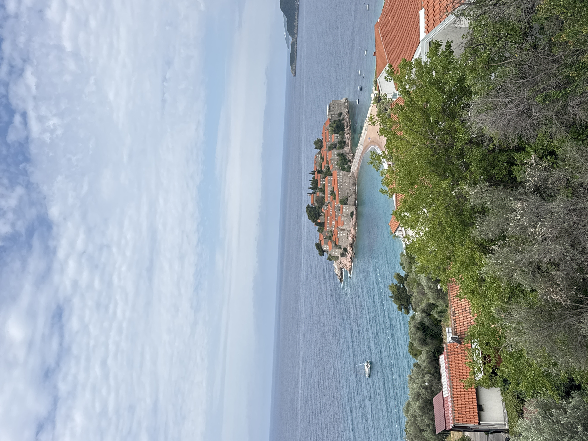 The image displays a captivating coastal landscape centered on Sveti Stefan, a small island settlement in Montenegro, viewed from an elevated perspective on the mainland. The island, densely covered with numerous stone buildings featuring distinct red-tiled roofs, is connected to the shore by a narrow, sandy causeway. Lush green trees and vegetation are interspersed among the buildings on the island and along the adjacent mainland.
The clear, deep blue waters of the Adriatic Sea surround the island and stretch towards the horizon, displaying gentle ripples. Several small boats are visible in the water, scattered both near the island and further out to sea. On the far left horizon, a distant landmass, likely part of the Montenegrin mountains or coastline, is subtly visible.
The sky above is a blend of light blue and white, with scattered, textured clouds suggesting an overcast yet bright daytime. The foreground on the right reveals more of the mainland, characterized by additional buildings with red-tiled roofs nestled within abundant green foliage. The scene captures the picturesque charm and tranquility of this famous Montenegrin location. No visible text is present in the image.