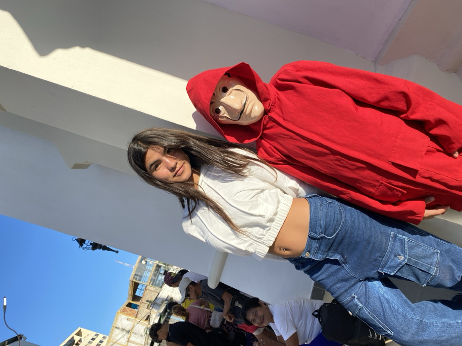 A young woman poses next to a mannequin in a red jumpsuit and a Salvador Dalí mask. She wears a white crop top hoodie and blue cargo jeans, revealing her navel piercing. The scene is outdoors, with a white building structure behind them. Other people are visible in the background, along with a building under construction. The sky is clear and blue. The photo has a slightly tilted angle.