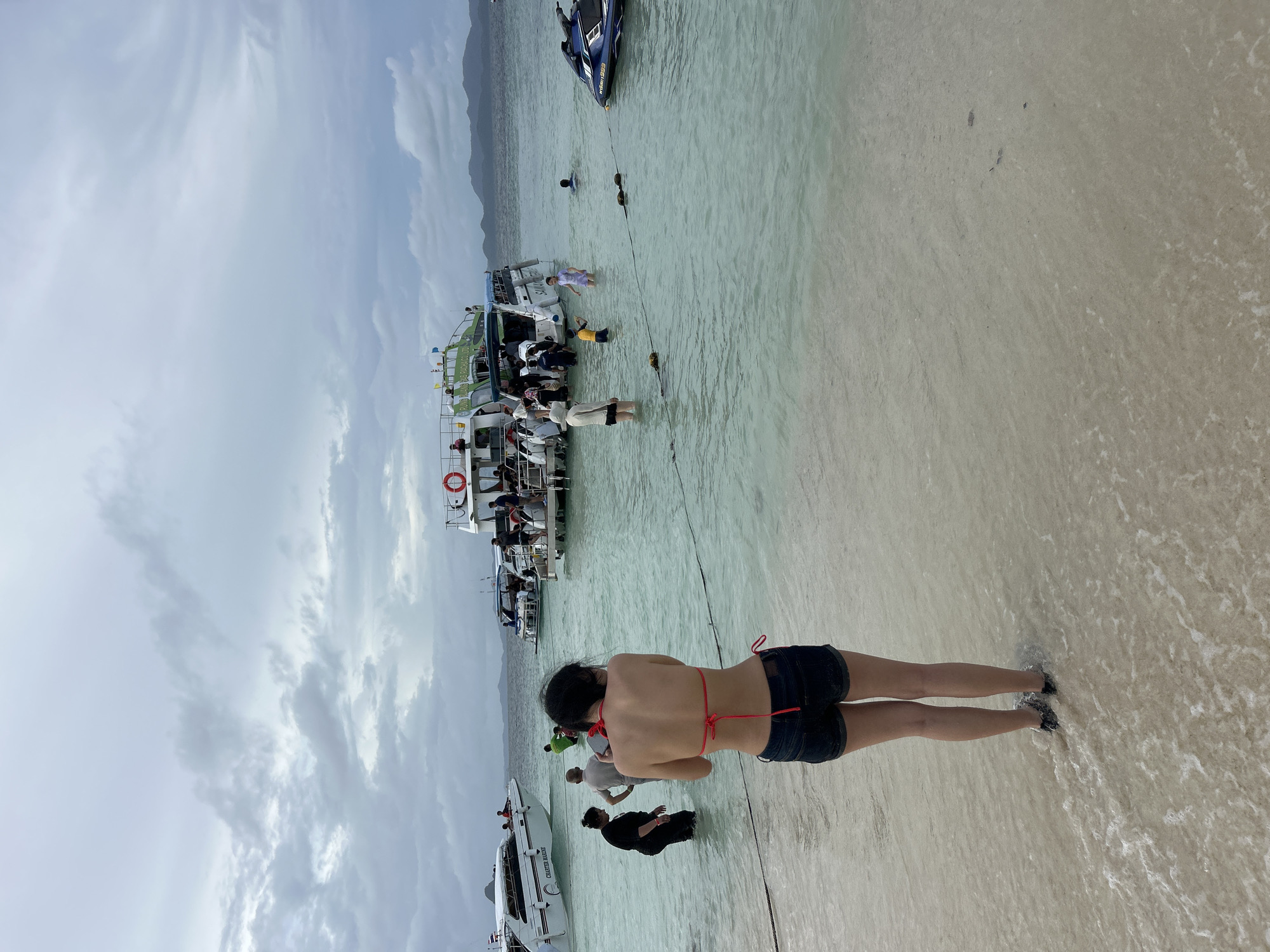 The image depicts a crowded beach scene in Tambon Phru Nai, Thailand. The water is clear and shallow, with several boats anchored offshore, including speedboats and a larger passenger boat. Many people are in the water, some wading and others swimming. On the beach, a woman in a bikini and denim shorts is facing away from the camera, looking out at the water. In the background, a jet ski is visible near the shore. The sky is overcast with scattered clouds, suggesting a moderate weather day. The overall impression is one of a popular tourist destination with active water-based recreation.