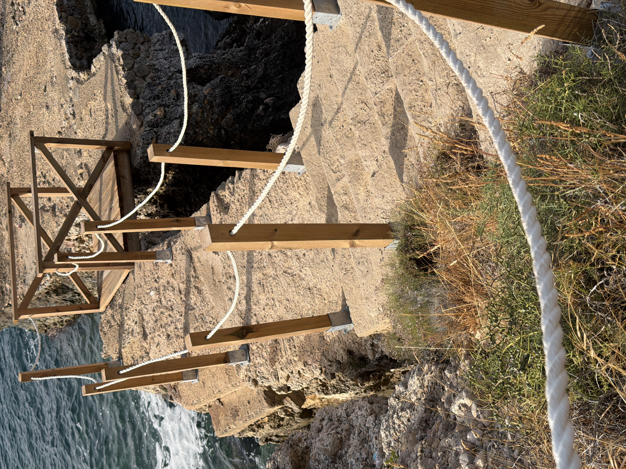 The image depicts a stone staircase leading down towards the sea in Calvià, Spain. The staircase is flanked by wooden posts and a white rope railing, designed to assist visitors navigating the rocky terrain. The sea, with its blue-green hue and gentle waves, is visible on the left side of the frame. The steps themselves appear weathered and worn, suggesting they have been in place for some time. Vegetation, including dry grasses and some green, spiky plants, grows along the sides of the path and between the rocks. In the background, a wooden walkway or platform is visible, constructed over the rocky coastline. The lighting suggests it is daytime, with shadows cast by the railing posts. The scene conveys a sense of natural beauty and a well-maintained pathway for accessing the coastal area.