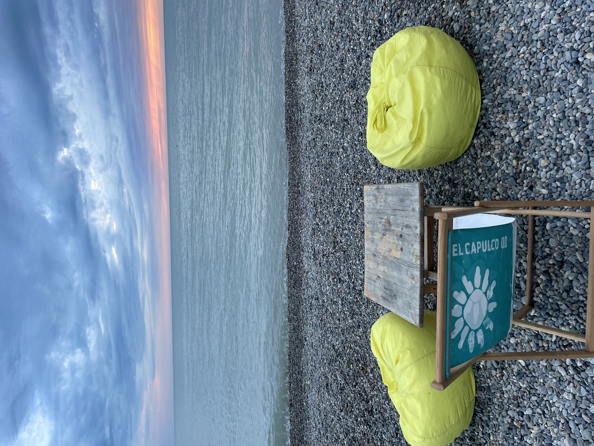 The image shows a beach scene in Sochi, Russia, featuring two yellow beanbag chairs, a small wooden table, and a chair with a turquoise back that reads "El Capulco 00" with a white sun-like design. These items are situated on a pebble beach next to the sea. The sky transitions from blue with clouds to a soft orange hue near the horizon, likely indicating sunset or sunrise. The water is calm, with gentle waves lapping at the shore. The scene suggests a relaxed, possibly private, beach setting.