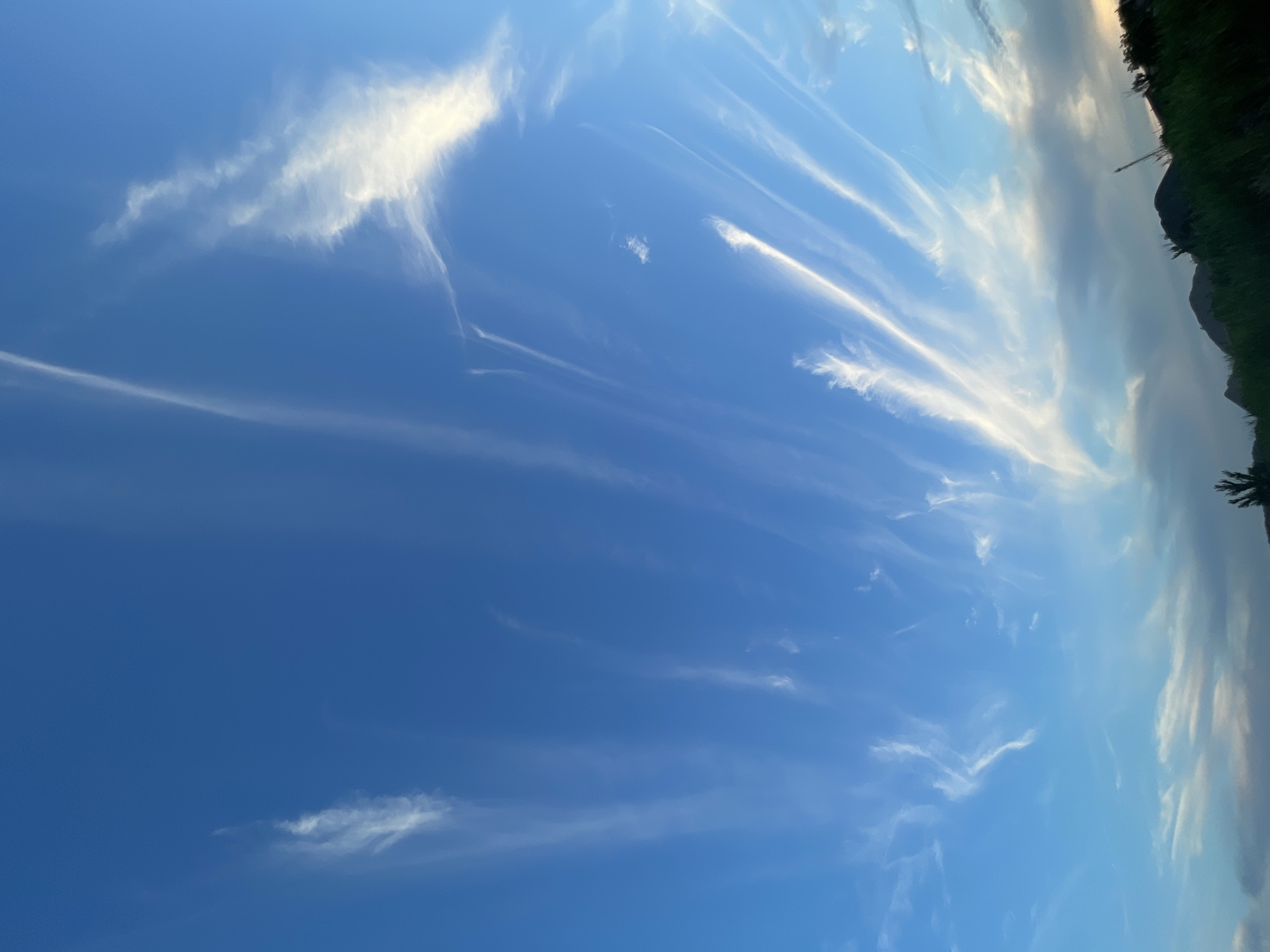 The image depicts a clear blue sky with wispy, white cirrus clouds stretching across it. The clouds are elongated and appear to be blown by the wind, creating a dynamic and streaky pattern. The lower portion of the image shows a silhouetted landscape with dark green grass and the dark shapes of hills or mountains. The overall scene suggests an outdoor environment, possibly a rural or semi-rural setting, under a bright sky. There are no people or specific man-made structures visible in the image that would indicate a precise location within Manhush, Ukraine, or any other city.