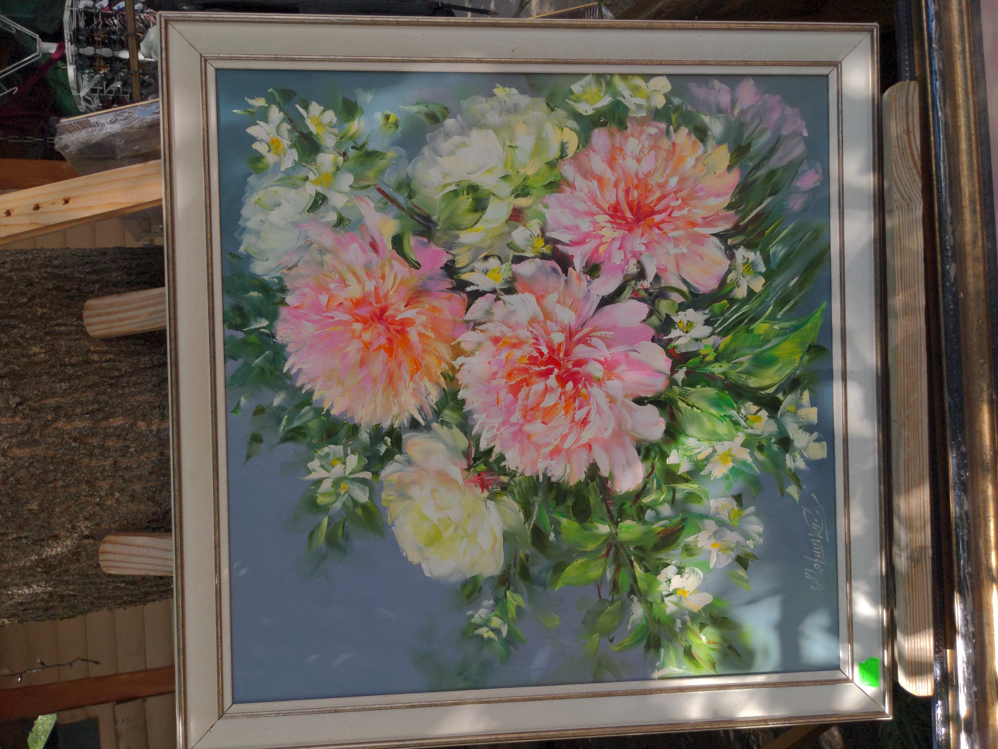A framed oil painting of a bouquet of peonies is displayed outdoors, leaning against a rough tree trunk. The painting features large, lush peonies in shades of pink, peach, and white, interspersed with smaller white flowers and green foliage. The background of the painting is a soft, muted blue. A signature, "Moshura," is visible in the lower right corner of the painting. The painting is framed in a wide, off-white frame with a gold inner bevel. A small, bright green sticker is affixed to the bottom left corner of the frame. The painting is set up on an easel, with wooden supports visible at the bottom. In the background, out of focus, are more wooden structures and possibly other items for sale. The scene appears to be at an outdoor market or exhibition. The location is the city of Holubyne, Ukraine.