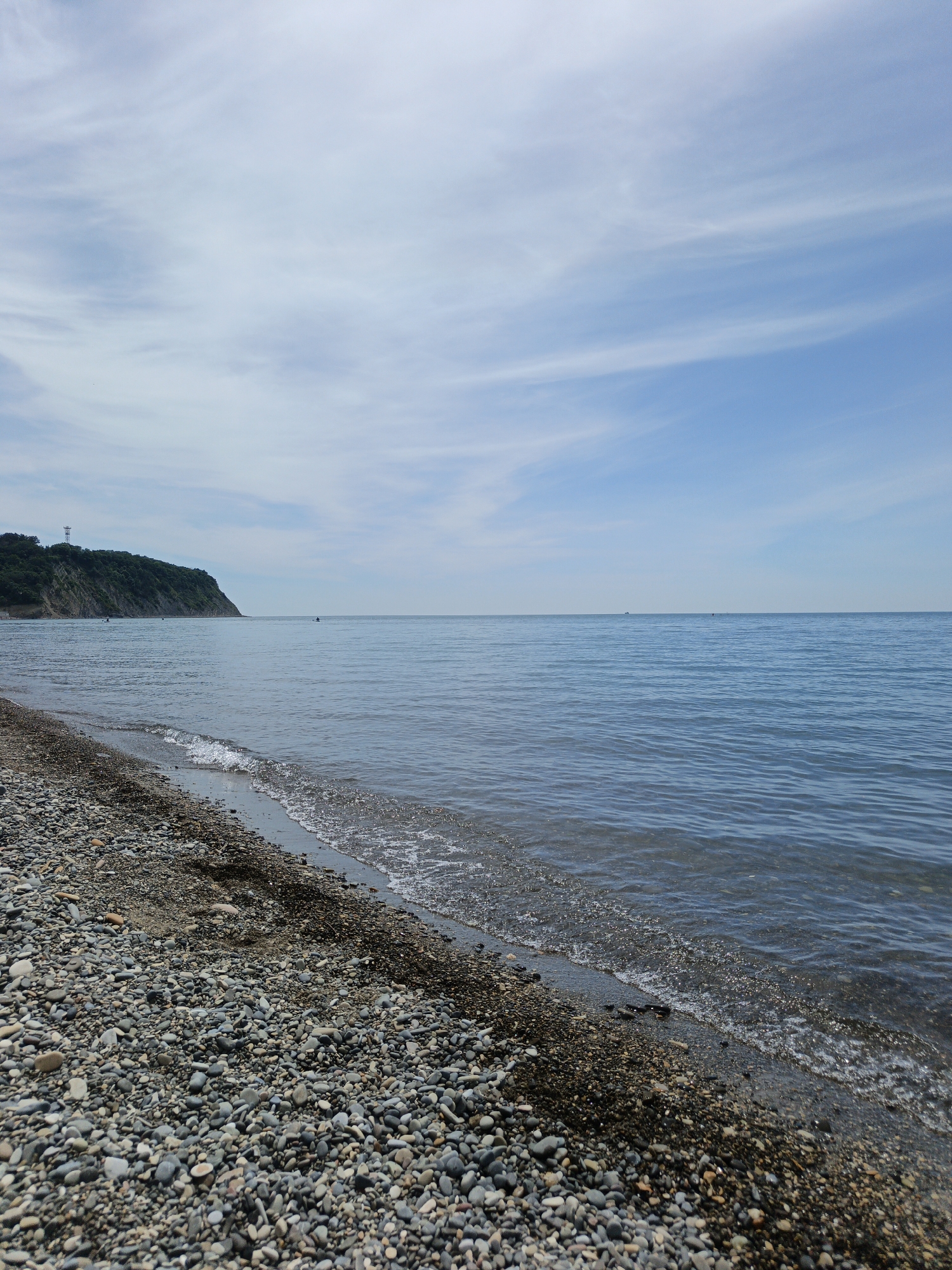 This image captures a tranquil coastal scene in Krinitsa, Russia, featuring a pebble beach, calm sea, and distant cliff under a partly cloudy sky. The upper two-thirds of the frame are dominated by a light blue sky streaked with wispy white clouds. A vast expanse of calm, deep blue-grey water occupies the mid-ground, meeting a clear, flat horizon line. Two small, dark vessels, likely kayaks or small boats, each with indistinct occupants, are visible on the water; one is positioned slightly left of center, and another smaller one is further to the right. A faint, larger vessel is barely discernible on the far left horizon.
To the left, a prominent, steep cliff covered with dense dark green vegetation extends into the sea. A tall, slender, light-colored tower stands atop this cliff. The foreground is composed of a pebble beach, stretching from the bottom left towards the center. The pebbles vary in size and color, predominantly shades of grey, brown, and light beige. Gentle waves with white foam break along the water's edge, wetting the stones and making them appear darker. Dark organic debris, possibly seaweed, is interspersed among the pebbles at the waterline. The image is captured from a low perspective on the beach, looking straight out towards the sea. No distinct human figures are visible on the beach or cliff.