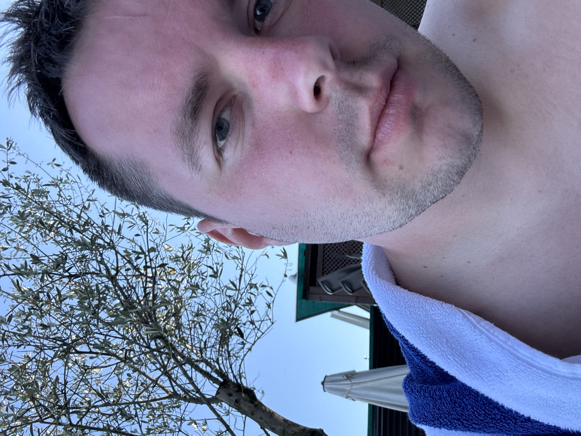 A close-up, low-angle selfie shows a fair-skinned man with short, dark hair and a light stubble beard. He is wearing a blue and white towel draped over his shoulders. His skin appears flushed, suggesting sun exposure. In the background, a clear blue sky is visible, with the branches of a tree extending from the upper left corner. Partially visible structures, including a green awning and white umbrellas, are in the middle ground. The setting appears to be an outdoor area, possibly near a pool or resort, in the Prince Islands, Turkey.
