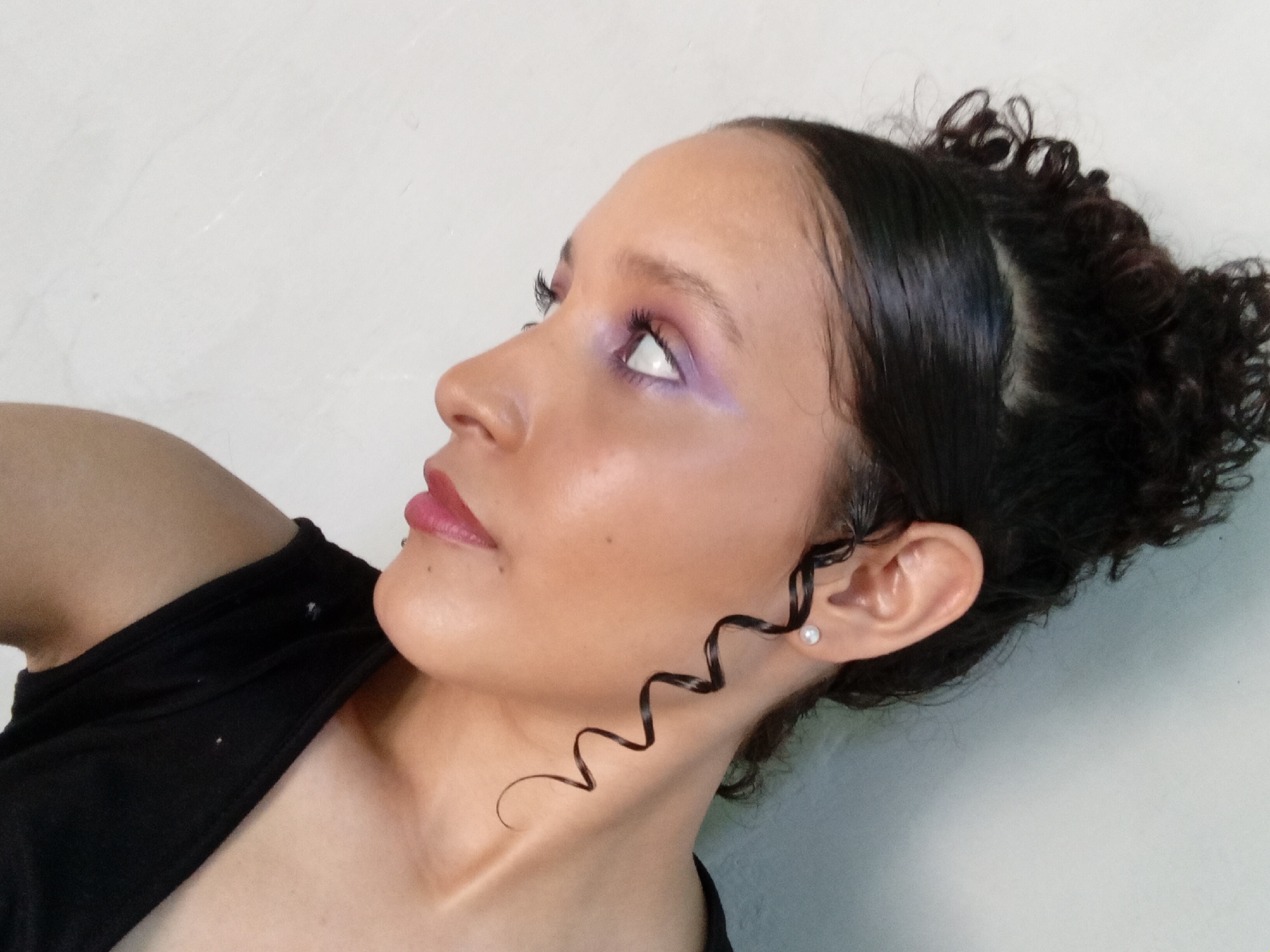 A woman with dark hair pulled back into a bun, with a curly strand framing her face, is looking upwards and to the left. She is wearing lavender eye makeup and lipstick. A pearl earring is visible in her left ear. She is wearing a black top. The background is a plain, light-colored wall. The image does not provide sufficient information to determine the location is Parroquia Ocumare, Venezuela.