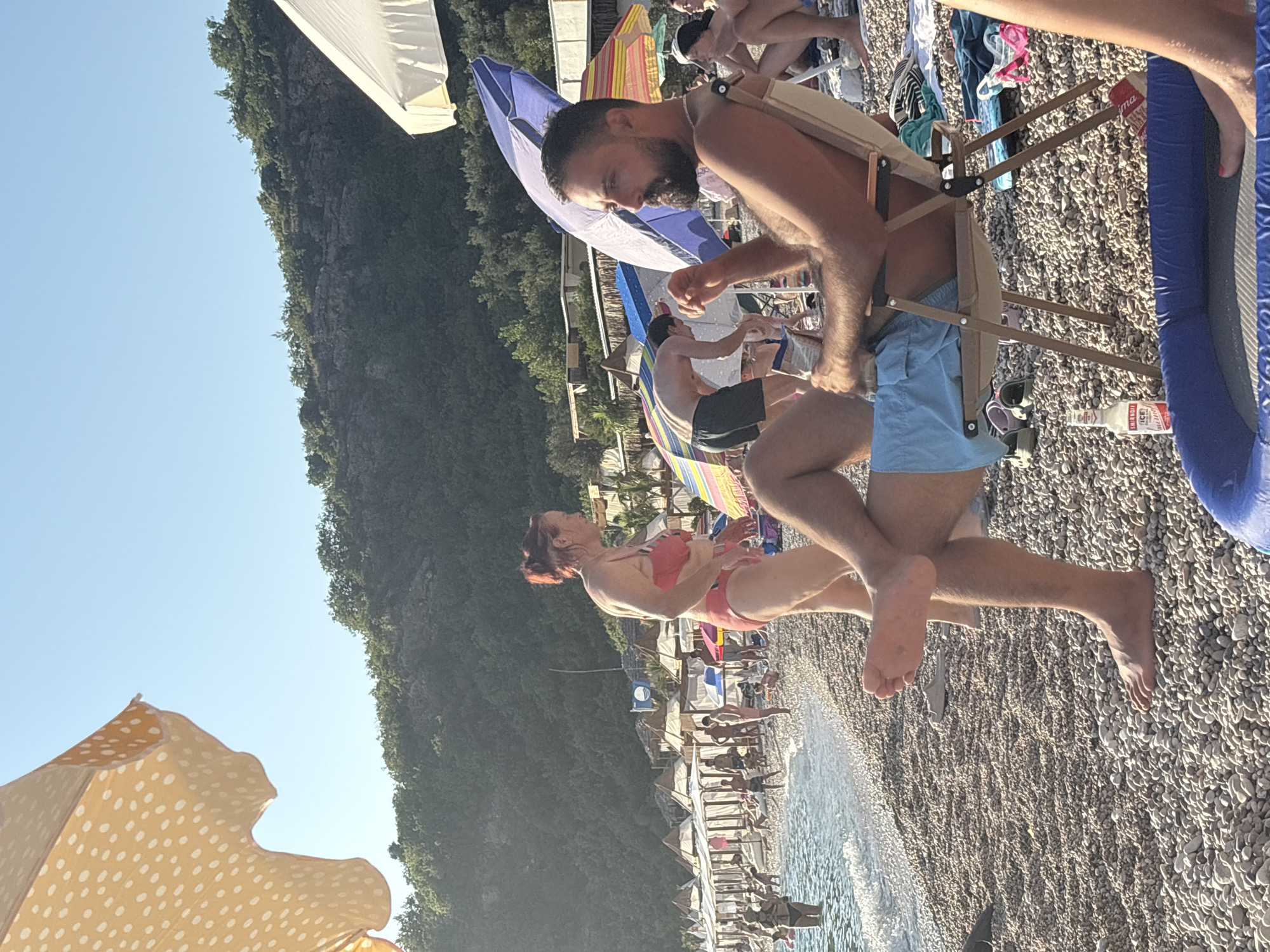 In the city of Drobnići, Montenegro, a man with a beard is seated in a beach chair on a pebble beach, facing right, with his legs crossed and wearing blue swim trunks. He is holding a white package. Behind him, a blue and white umbrella is positioned. To the left of the man, a woman in a red bikini stands, facing the sea. In the background, the sea with gentle waves is visible, with many people sunbathing and walking along the shore. Several beach umbrellas, some striped and some solid colored, are set up along the beach. Further back, a line of white beach tents with thatched roofs runs along the shore, and a forested hillside rises behind the beach. The sky is clear and blue.