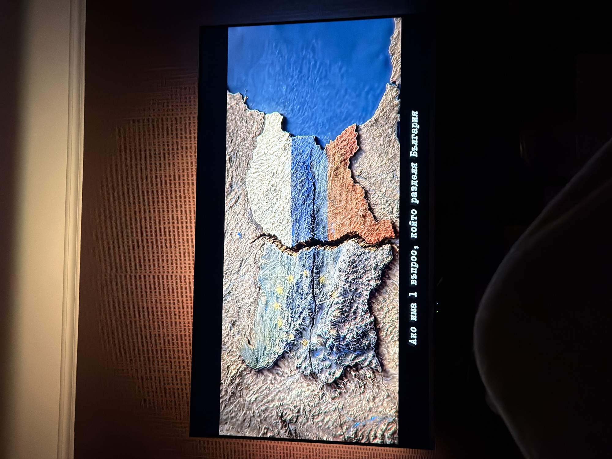 The image shows a screen displaying a relief map of Bulgaria. The northern part of the map is colored white and blue, the eastern part orange, and the southern part is colored to mimic the EU flag (blue with yellow stars). A crack runs horizontally across the map, dividing it in two. To the right of the map, text in Bulgarian reads "Ако има 1 въпрос, който разделя България" (If there is one question that divides Bulgaria). The screen is dark, and the environment surrounding it is dimly lit, with a patterned wall visible to the left of the screen. The partial form of a person's silhouette is visible to the right of the screen.