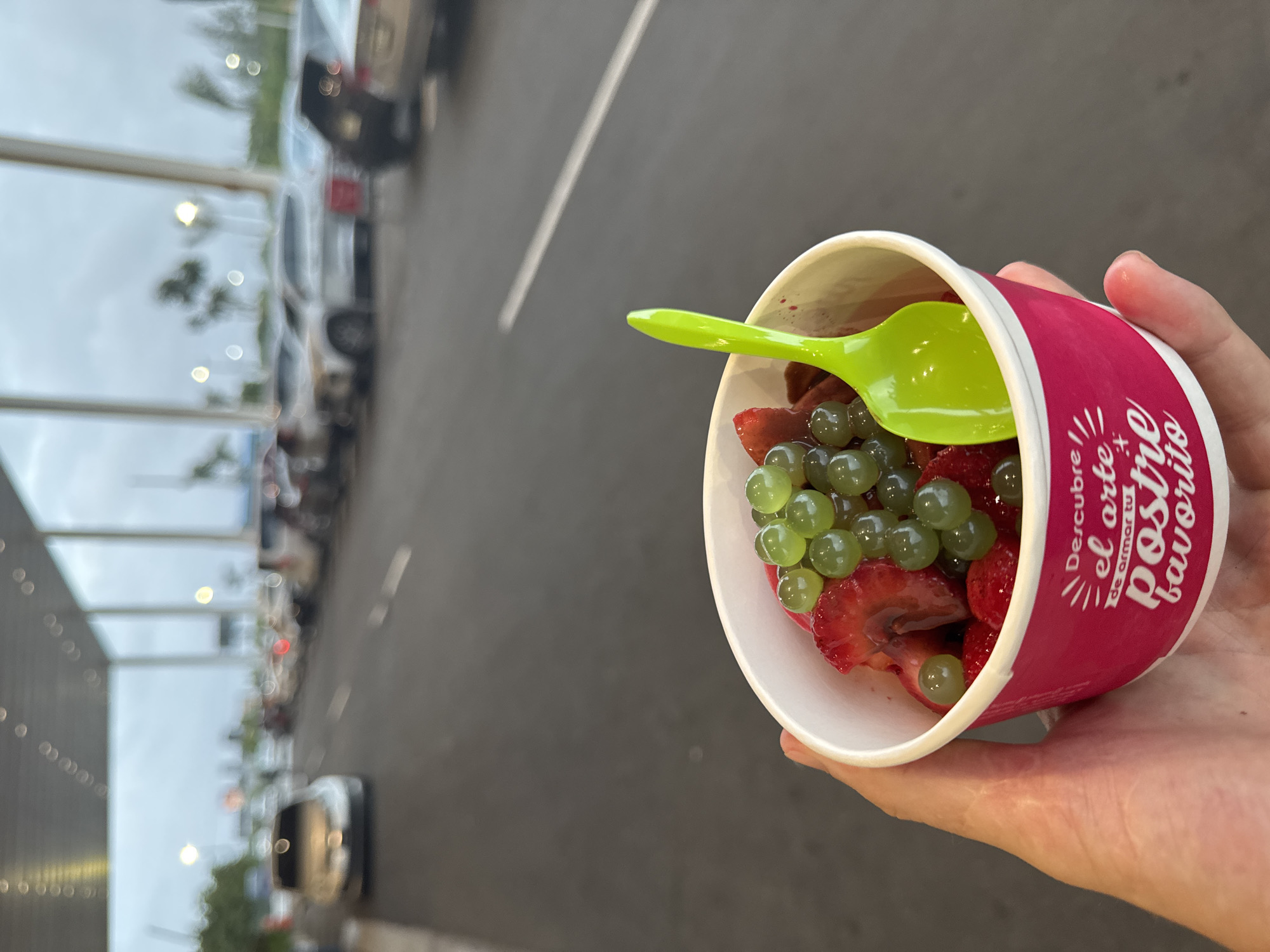 A person's hand holds a white paper cup filled with frozen yogurt, strawberries, and green boba pearls. A bright green plastic spoon is inserted into the cup. The cup has pink text that reads "Descubre el arte de armar tu postre favorito". In the background, cars are parked in a row along a grey asphalt road, with a covered walkway structure overhead and a cloudy sky. This scene appears to be in the parking area of a commercial establishment in Higüey, Dominican Republic.