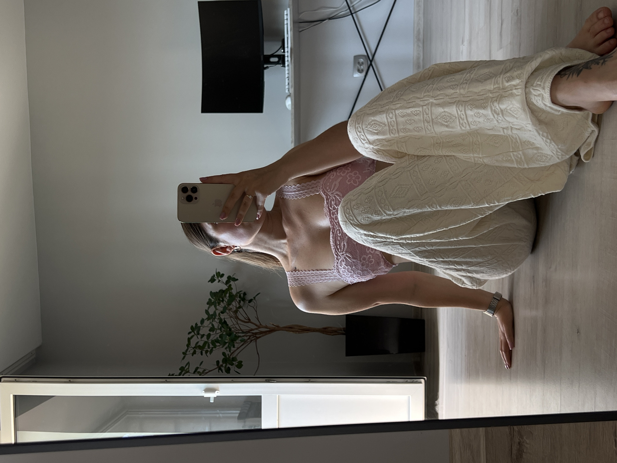 A young woman, facing a mirror and taking a selfie with her smartphone, is sitting on a light-colored wooden floor. She is wearing a pink lace bralette and loose-fitting, cream-colored textured pants. Her left hand rests on the floor, and she is wearing a silver watch. Behind her, a tall potted plant with green leaves is visible. In the background, a desk with a computer monitor and keyboard is set up against a grey wall. A large mirror to the left of the frame reflects the scene. The overall setting appears to be an indoor space, likely a room within an apartment in Ternopil, Ukraine, given the user's instructions, though specific identifying markers of Ternopil are not present in the image itself. The lighting suggests it is daytime, with natural light coming from a window or door on the left.
