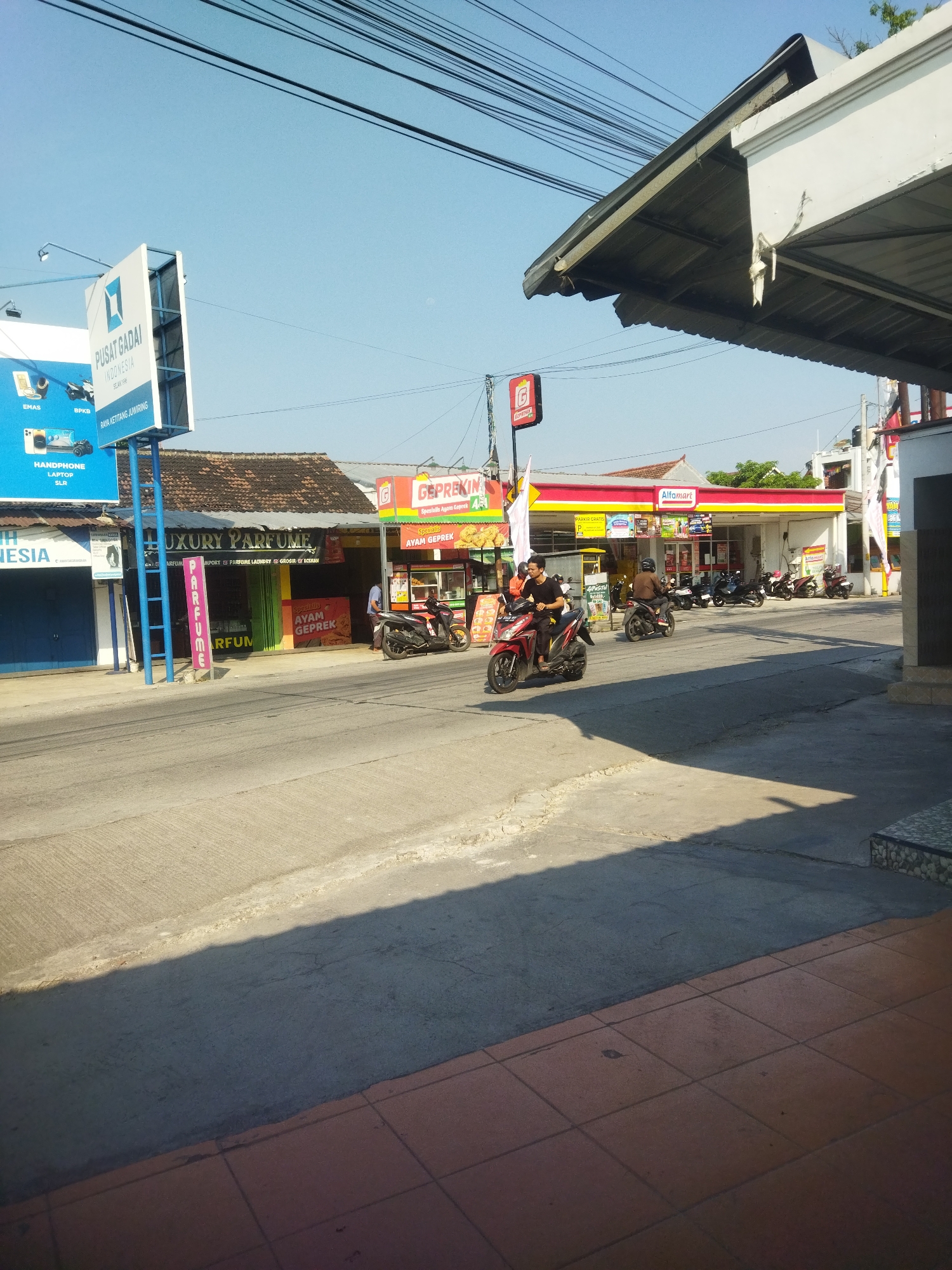 This is a daytime, eye-level, outdoor photograph taken on a street in Juwiring, Indonesia, showing a commercial area. The scene is dominated by a street in the foreground and a row of buildings and a gas station on the right side. The sky is clear and blue.
In the foreground, a paved street with a textured surface occupies the lower half of the image. A distinct shadow from an overhead structure falls across the street from right to left. On the far right, a sidewalk with reddish-brown square tiles is visible.
On the left side of the street, there is a multi-story building with blue and white signage. One sign, positioned high up, has the words "PUSAT GADAI INDONESIA" and "SELAMAT DATANG" written on it, along with an image of a blue diamond logo. Below this, there are smaller signs advertising "HANDPHONE", "LAPTOP", and "SLR". Next to this building, another structure has a dark blue facade and a sign that says "LUXURY PARFUME". Adjacent to "LUXURY PARFUME" is a shop with a green and yellow striped facade, and a sign that reads "PARFUME". Further to the right, there is a storefront with a red and yellow sign displaying "AYAM GEPREK".
On the right side of the street, a brightly lit building with a red and yellow awning houses an "Alfamart" convenience store. Several motorcycles are parked in front of the store. A red sign with a white 'G' is visible on a pole near the Alfamart. Further down the street, there are more buildings and parked motorcycles.
There are several people visible in the image. Two individuals are riding motorcycles on the street. The person on the red motorcycle in the mid-ground is wearing a black t-shirt. The person on the brown motorcycle behind them is wearing a helmet. Another person, possibly an employee, is standing outside the "AYAM GEPREK" shop on the left.
The environment appears to be a busy street with shops and a gas station, typical of a commercial district. Overhead power lines are visible against the sky. The lighting suggests it is daytime, with the sun casting strong shadows.