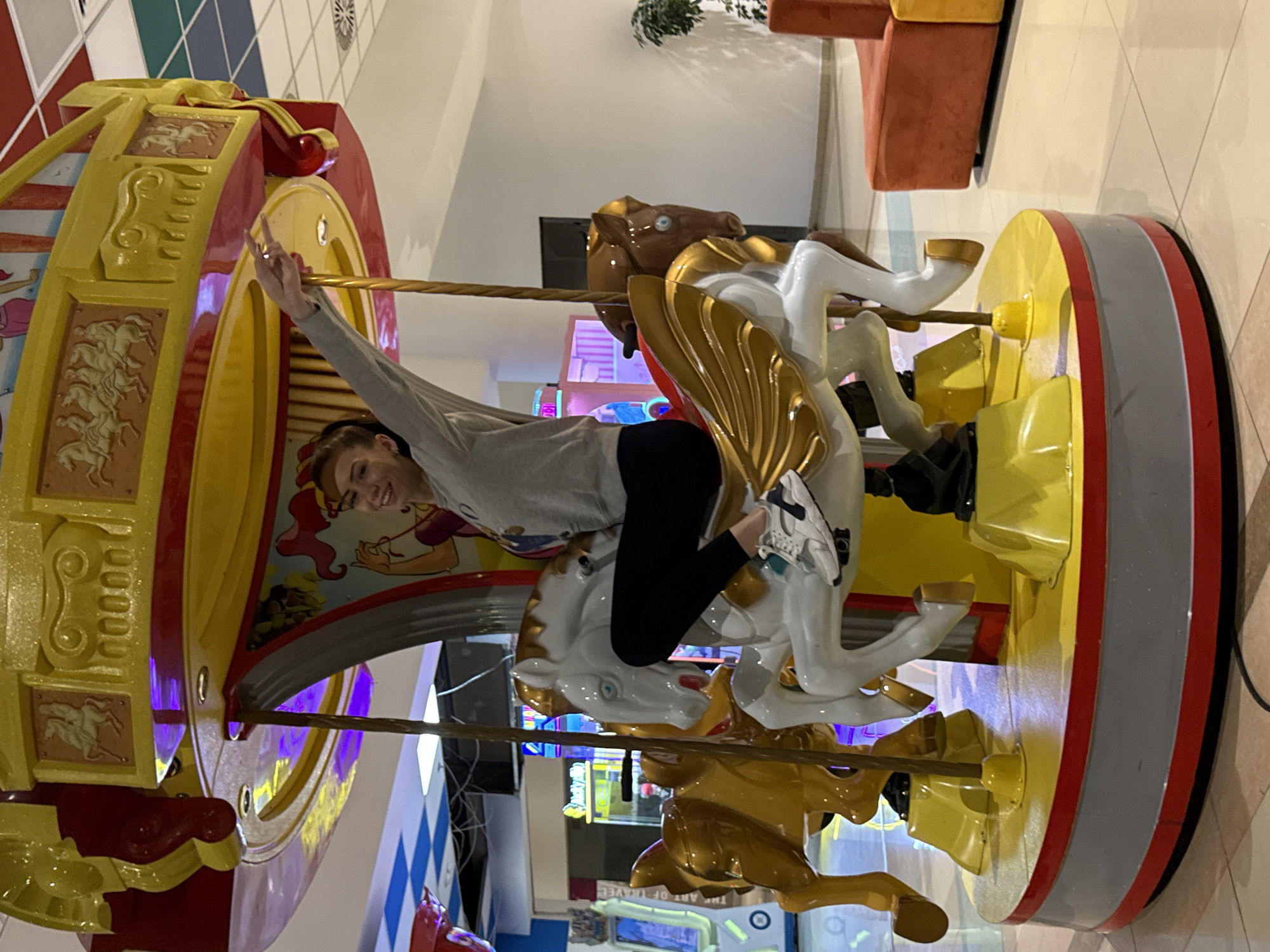 The image depicts a young woman riding a carousel in an unknown city and location. The carousel is decorated with ornate horses and colorful panels. The woman is wearing a gray sweater and black pants. She has one arm raised in the air. The surroundings suggest the carousel is indoors, possibly in a shopping mall or entertainment center. The floor is tiled, and there is some furniture visible in the background.