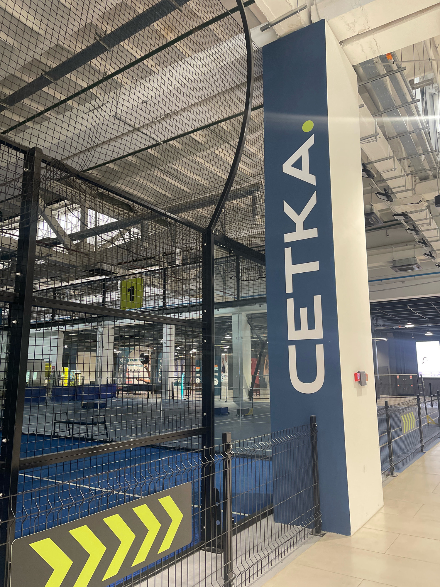 The image shows the interior of an indoor sports facility, possibly a padel or tennis court, located in Orenburg, Russia. A large blue and white pillar with the word "CETKA" in white capital letters and a green dot is prominently displayed on the right side of the frame. The pillar is adjacent to a dark metal fence that encloses a blue sports court with white markings. Above the court, a black mesh netting system covers the ceiling. In the background, other sports courts and the facility's infrastructure, including lighting and ventilation systems, are visible. A person can be seen in the background on one of the courts, holding a paddle. A sign with yellow arrows is attached to the fence in the foreground.