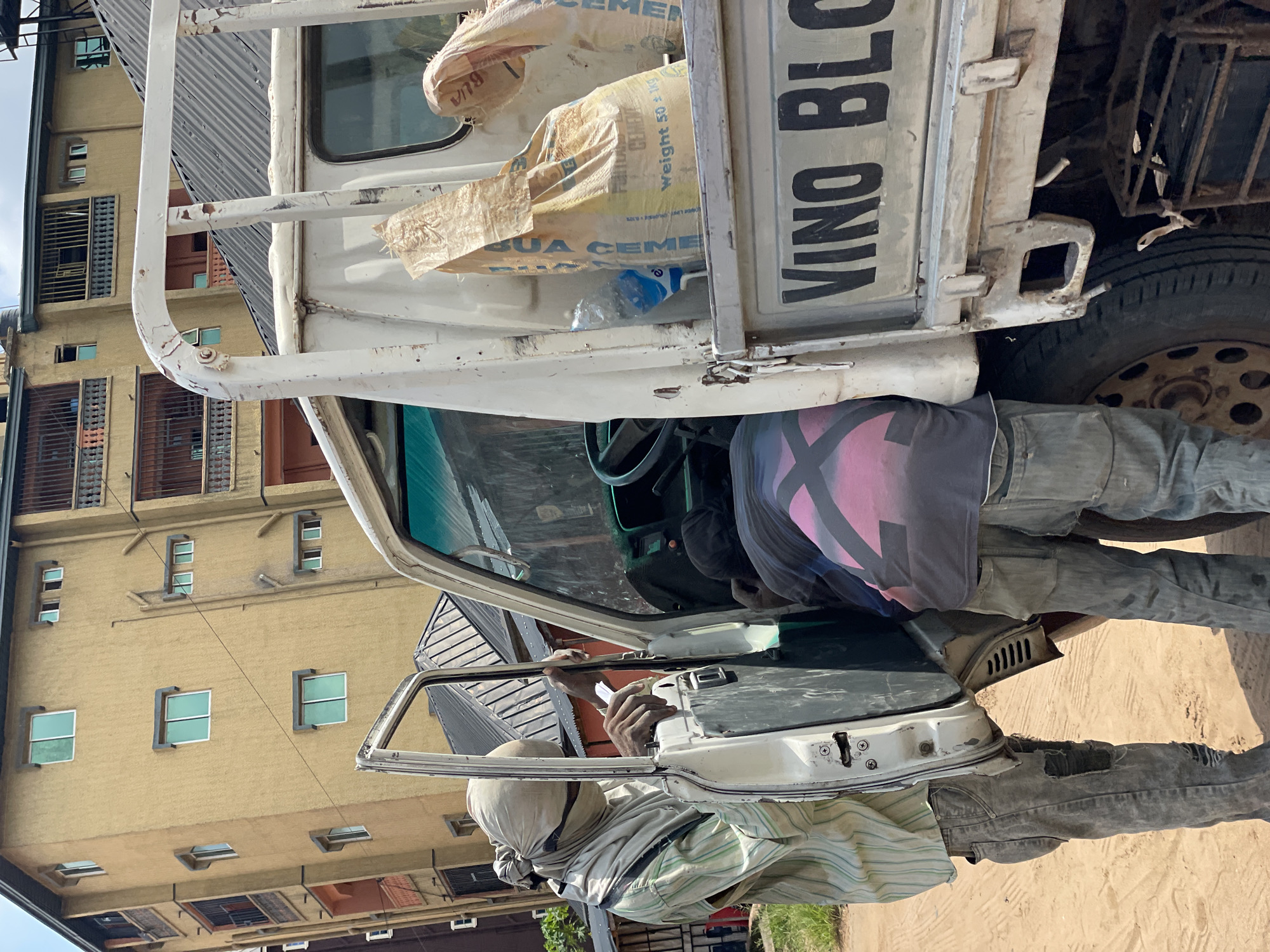 In a dusty, outdoor urban environment, two individuals are interacting with the open driver's side door of a white pickup truck. The person on the left is partially obscured, wearing a light-colored striped shirt and gray, ripped jeans. They have a light-colored cloth wrapped around their head and face. Their hands are visible, with one holding the edge of the truck door and the other holding a white object, possibly paper. The second person is leaning into the truck cab from the right side, wearing a dark t-shirt with a pink and black geometric design and gray jeans. The truck bed contains two large sacks of what appears to be cement, one with visible text indicating "Weight 50 + 1kg," and a partially visible plastic water bottle. The side of the truck bed displays large black lettering that reads "VINO BLO." In the background, a multi-story building with yellow-toned walls and numerous windows dominates the scene. The ground is composed of dirt.