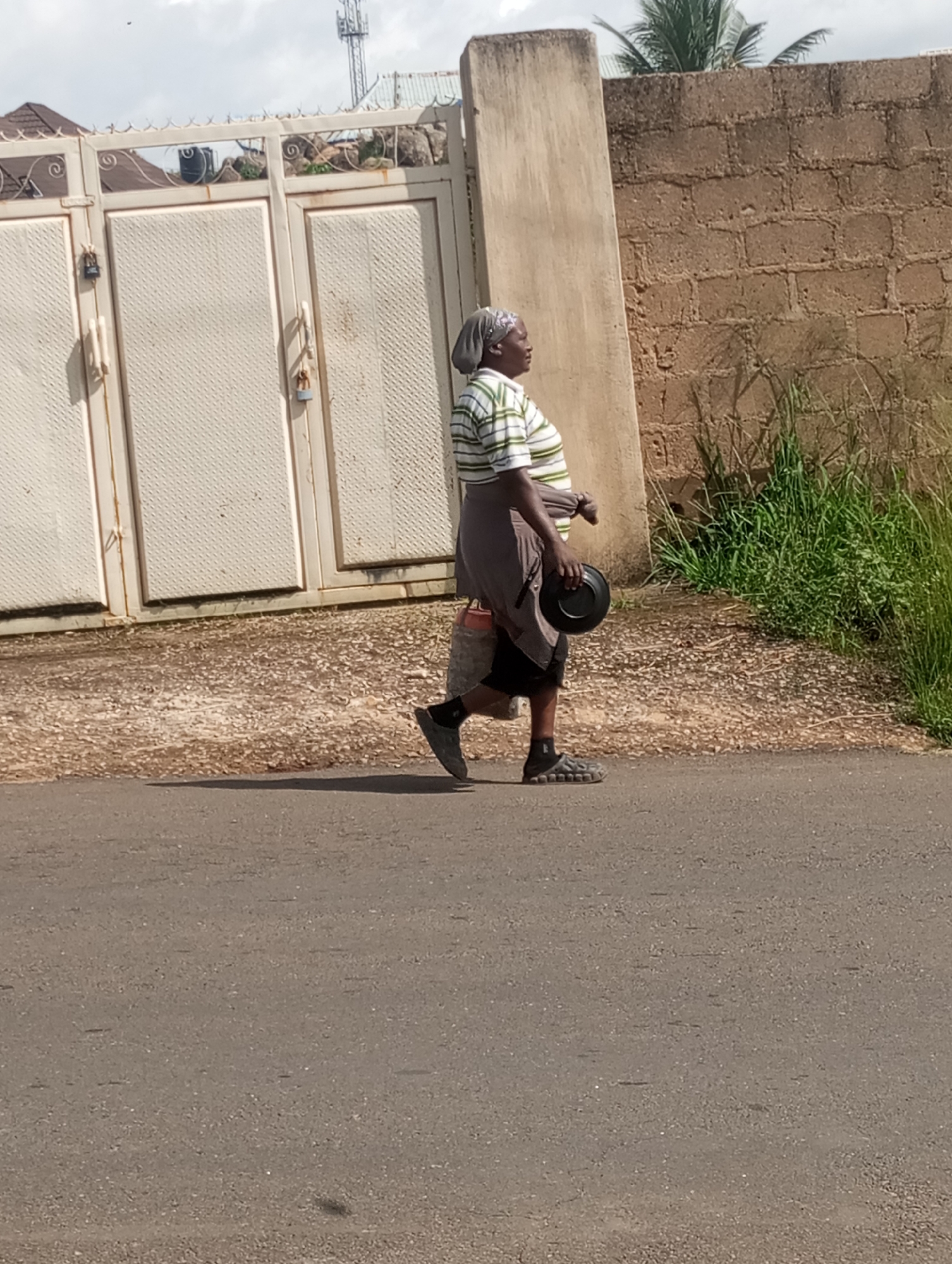 A woman walks on a paved road in Jos, Nigeria. She is wearing a striped t-shirt and a grey skirt, with a grey headscarf. She carries a black bowl in her right hand and a bag slung over her left shoulder. The road is bordered by a gravelly area and a brick wall. Behind the woman, a large metal gate is visible. A cell tower stands in the background against a cloudy sky. The overall scene depicts an ordinary day in a Nigerian city.
