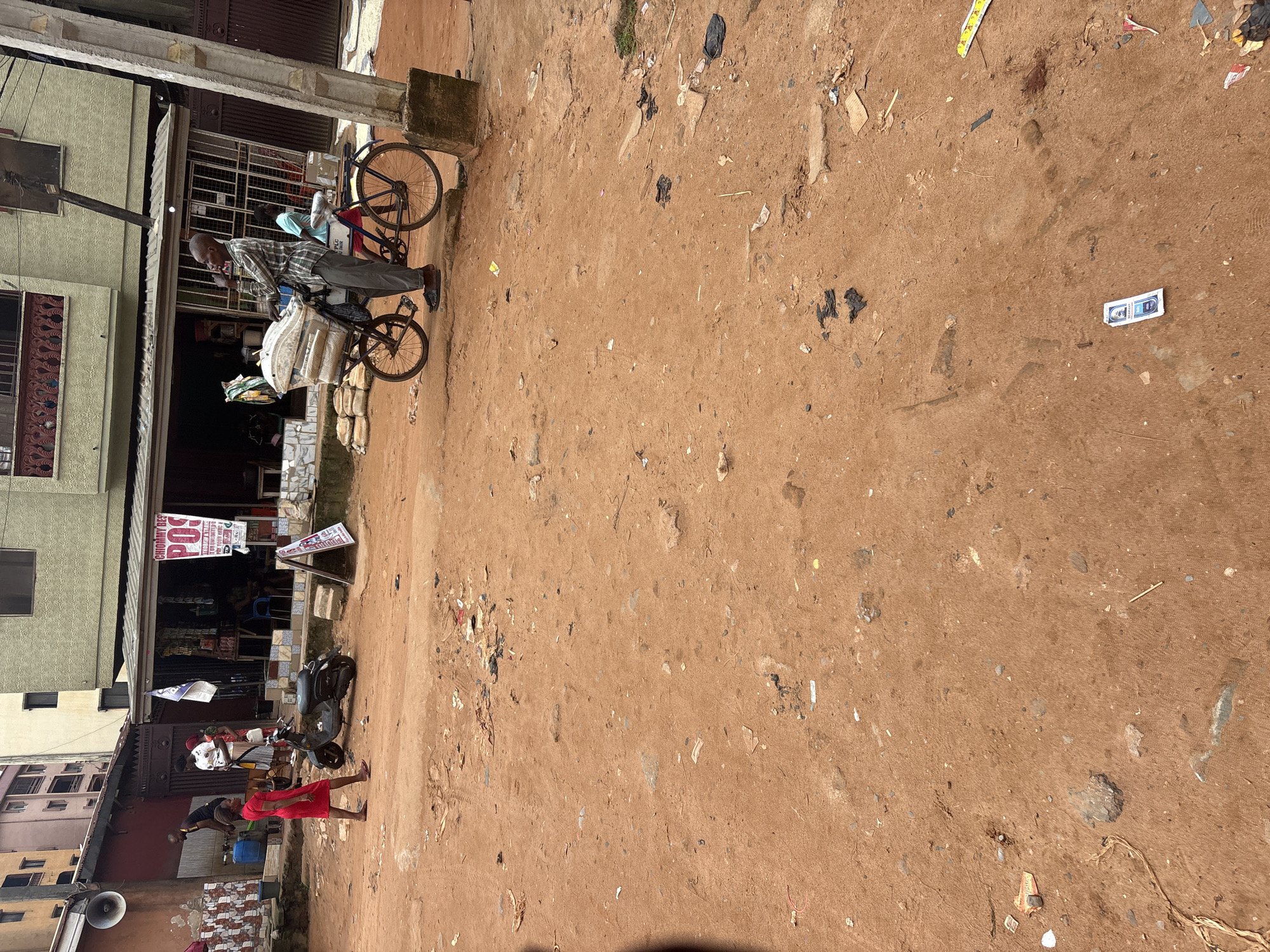 The image depicts a dusty street scene in Onitsha, Nigeria, likely near a commercial area. In the foreground, a woman in a red dress walks across the reddish-brown dirt road. A parked scooter is visible to her left. Further into the scene, a man in a plaid shirt and dark pants stands beside a bicycle loaded with sacks. He appears to be smoking and looking to the side. Behind him, a person in a light blue shirt is seated on the bicycle. The background shows a building with an open storefront, displaying various goods and a banner advertising "CHIOMMY BES POS." The ground is littered with debris, including plastic bags, wrappers, and other trash, indicative of a busy urban environment. A utility pole and a concrete pillar are also present on the right side of the frame. The overall impression is one of everyday street life in a Nigerian city.