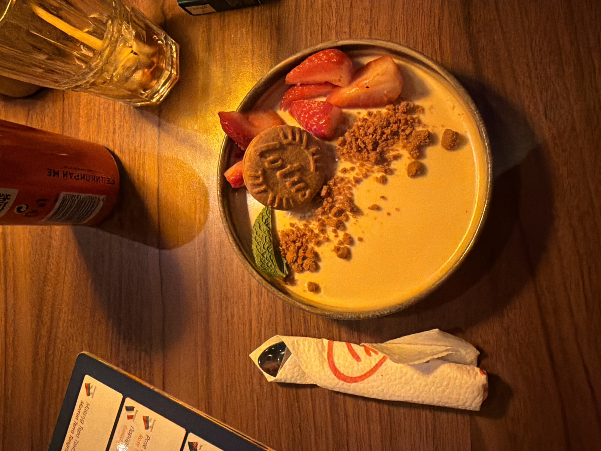 The image captures a dessert plate on a wooden table in what appears to be a restaurant or cafe. The plate contains a light yellow dessert with sliced strawberries, crumbled biscuit, and a single cookie prominently displayed. A mint sprig adorns the dessert. A glass of a beverage with slices of fruit (possibly lemon or orange) is partially visible at the top left, along with a can of a soft drink with Cyrillic text on the label. A wrapped utensil (likely a spoon) rests beside the plate. A menu with Cyrillic text is visible in the lower-left corner. The scene suggests the location is in Sofia, Bulgaria due to the Cyrillic text present on the menu and drink can. The objects are arranged on a wooden surface, likely a table in a dining establishment. There are no people visible in the frame. The scene suggests the environment is dimly lit.