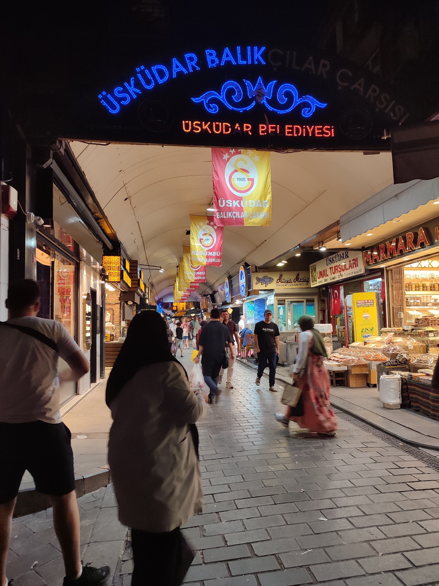 The image depicts a nighttime street scene in the Üsküdar Fish Market (Üsküdar Balıkçılar Çarşısı) in Üsküdar, Turkey. A prominent blue neon sign overhead reads "ÜSKÜDAR BALIKÇILAR ÇARŞISI" and a smaller red neon sign below says "ÜSKÜDAR BELEDİYESİ" (Üsküdar Municipality). Several banners hang from the arched ceiling of the market, featuring the Galatasaray football club logo and Turkish text.
The street is paved with stone. On the left side, shops line the market, with glass windows displaying merchandise and bright interior lighting. On the right side, market stalls are set up, displaying dried fruits, nuts, and other food items. People are walking along the market street. In the foreground, a person wearing a grey coat and a headscarf walks away from the camera. To their left, a person in a light-colored shirt and dark shorts is also walking away. Further down the market, other individuals are visible, some walking, some standing, and some interacting with vendors. The overall atmosphere is that of a bustling nighttime market.