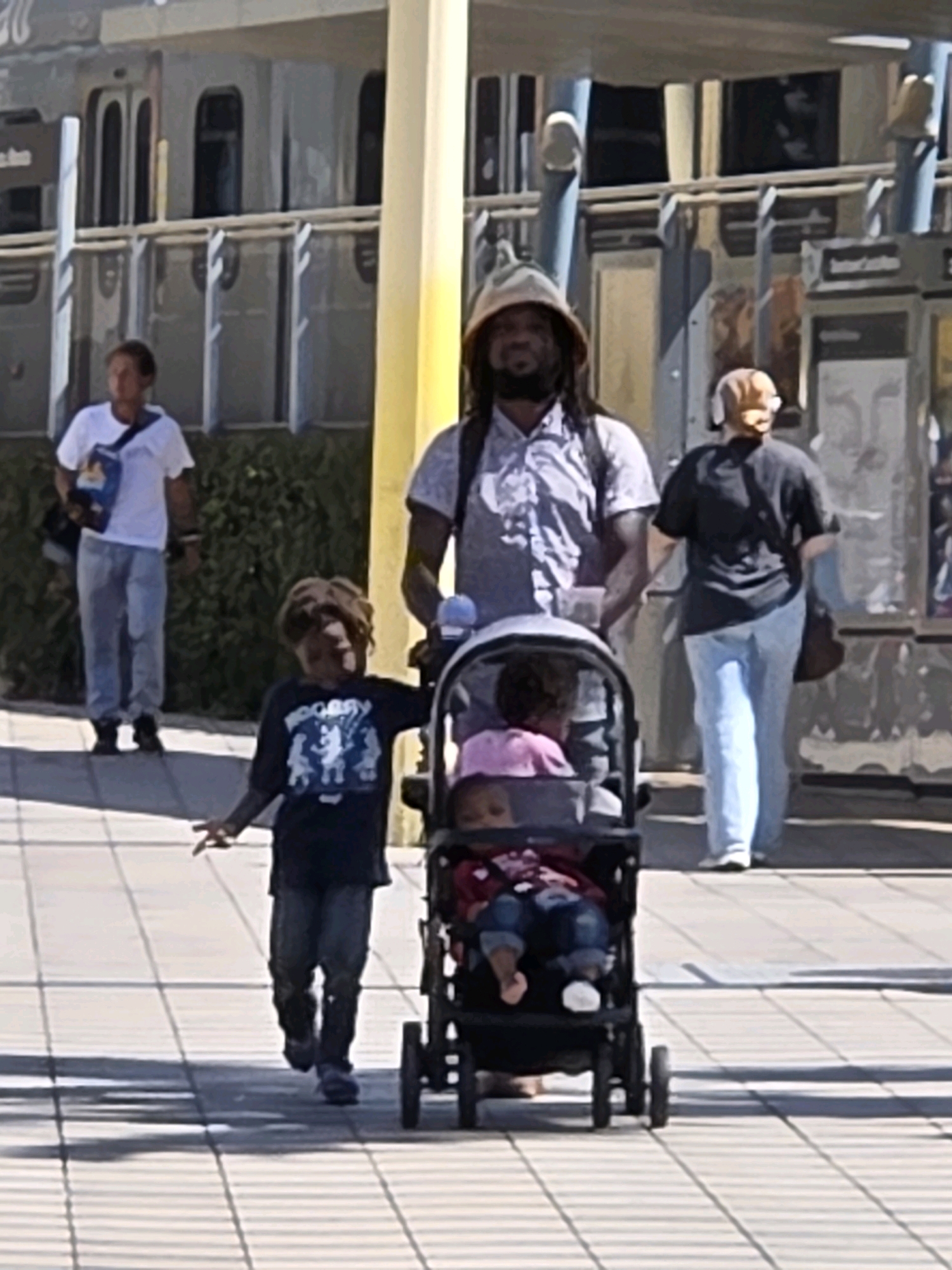 In Santa Monica, California, an adult male wearing a bucket hat and a patterned button-down shirt pushes a stroller with two young children. To his left, a young child in a dark t-shirt walks ahead, while a person in a white t-shirt and blue jeans stands further in the background. To the adult's right, another adult in a black t-shirt and blue jeans walks away from the camera. The scene takes place outdoors on a paved walkway with striped shadows, with a light-colored building and what appears to be a train station in the background.