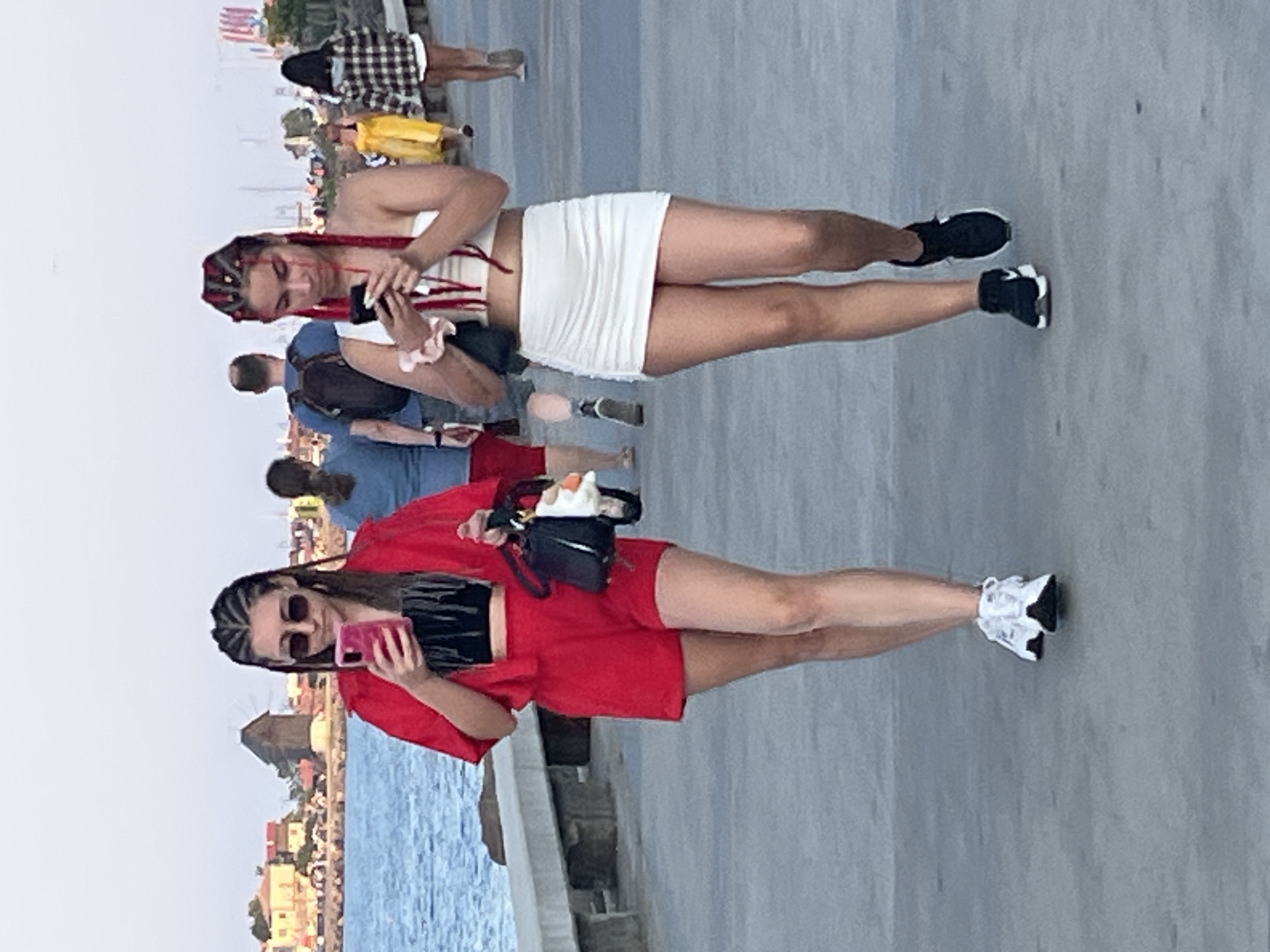 In Nesebar, Bulgaria, two young women with distinctive braided hairstyles are walking on a paved promenade. The woman on the left wears a red outfit consisting of a short-sleeved jacket and matching shorts, a black crop top, sunglasses, and white sneakers. She is holding a pink smartphone in her right hand and a black handbag with a white charm in her left. The woman on the right is dressed in a white tube top and a white ruched mini skirt, complemented by black sneakers with white soles. She is also looking at a smartphone in her hands. Several other people are visible in the background, including a man with a backpack, a woman in a blue shirt, a woman in a yellow dress, and a woman in a plaid jacket. The background features a body of water and buildings, suggesting a coastal urban setting.