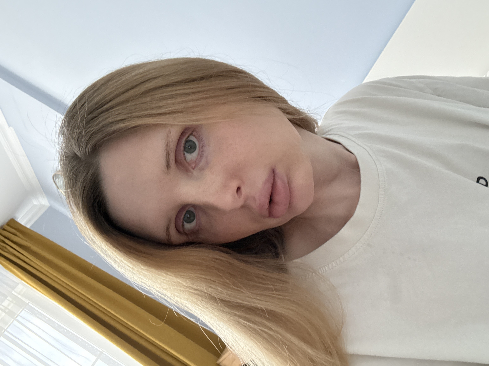 An overhead, close-up shot shows a woman with blonde hair and light skin. She is wearing a white t-shirt. Her eyes are open, and she is looking forward. Her lips are full. The background is a blue wall and a yellow curtain. The lighting is natural, coming from a window.