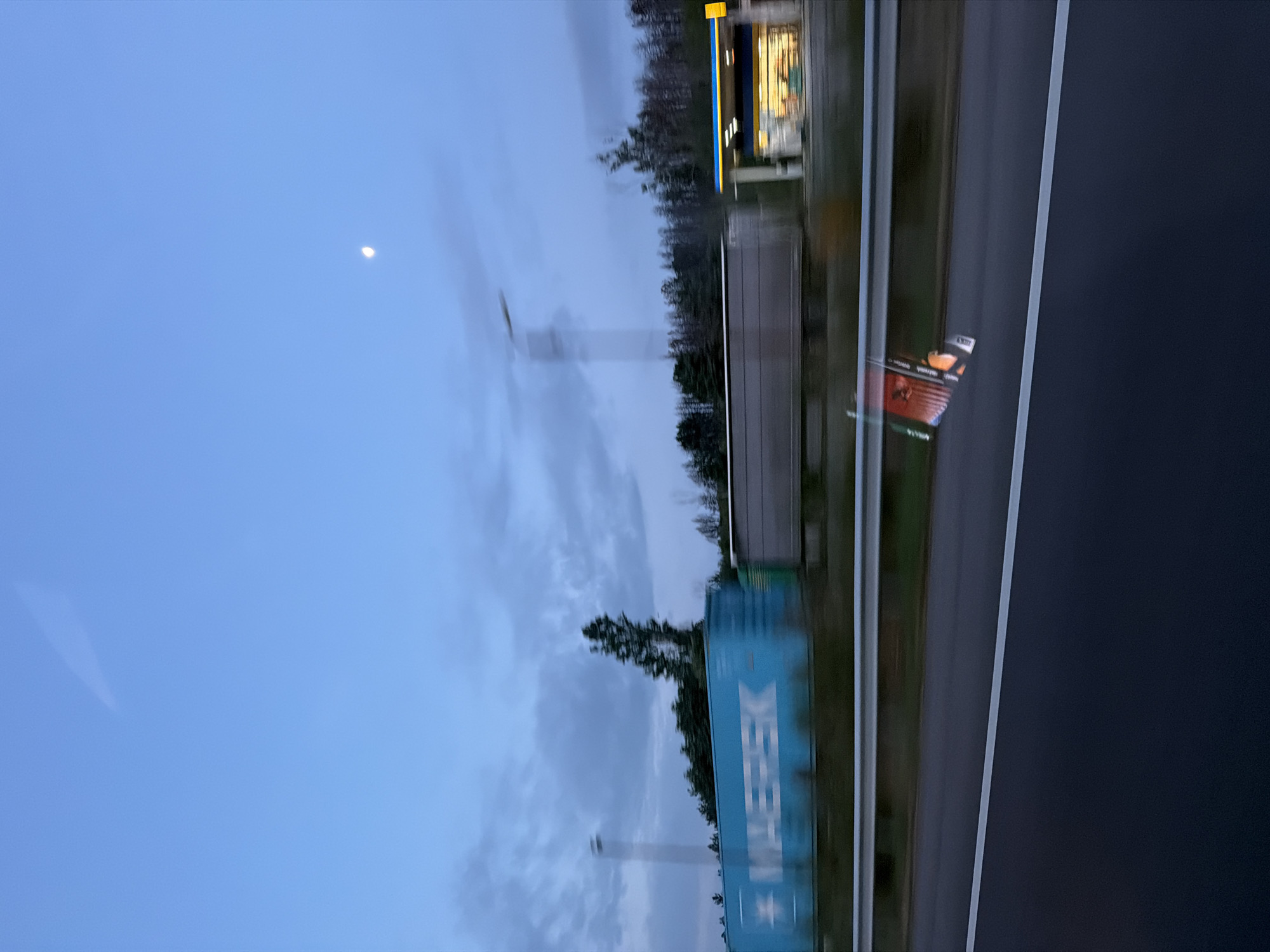This image captures a dynamic view from a moving vehicle, likely at dusk or dawn, given the deep blue sky and visible crescent moon. The scene is oriented vertically in the original, but describes a horizontal landscape. The blur in the foreground and mid-ground elements indicates significant motion.
In the foreground, a paved road with white lane markings is visible on the right, suggesting a highway or motorway. Along the left side of the road, the landscape features scattered trees and vegetation. A prominent blue shipping container with the word "MAERSK" clearly visible in white capital letters on its side dominates the lower mid-ground. Further down the road, a brightly lit building with distinctive yellow and blue architectural elements is seen, resembling a gas station or a roadside convenience store. A tall, slender structure, possibly a wind turbine or a transmission tower, pierces the sky in the distance. On the far right edge of the road, a small, orange and white reflective marker or sign is partially visible. The upper portion of the image is dominated by a clear blue sky with soft clouds and a distinct, luminous crescent moon.
The activity depicted is travel, viewed from inside a vehicle, possibly passing through or near Kalugerovo, Bulgaria. The time of day is characterized by low natural light, with artificial lights from the building standing out.